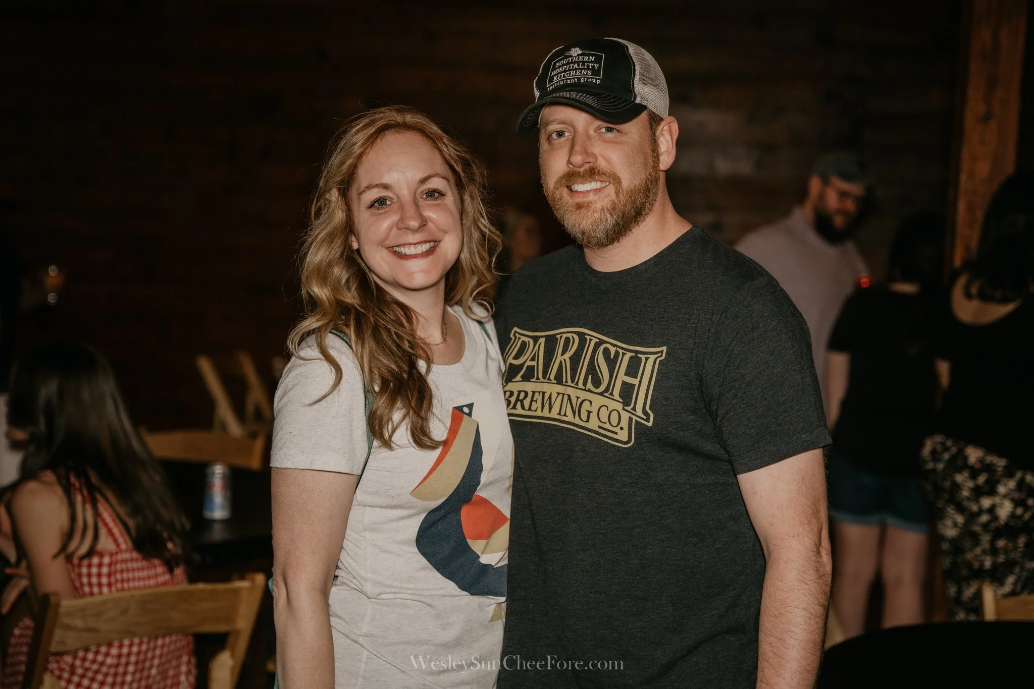 A smiling woman with wavy, shoulder-length red hair and a man with a beard wearing a black Parish Brewing Co. t-shirt and a cap, standing together in a dimly lit indoor venue with wooden walls, other people, and tables in the background.