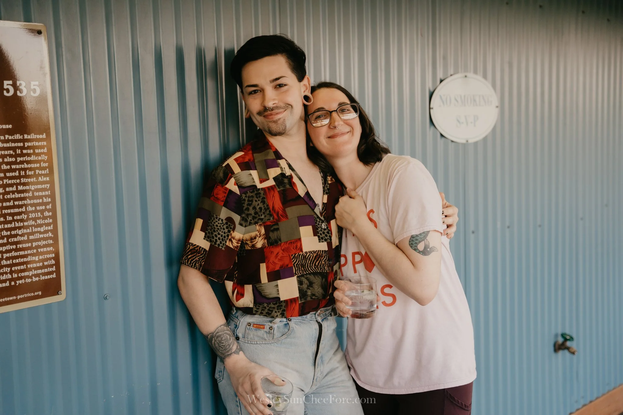 Two people smiling and hugging against a blue metal wall, holding drinks, with a 