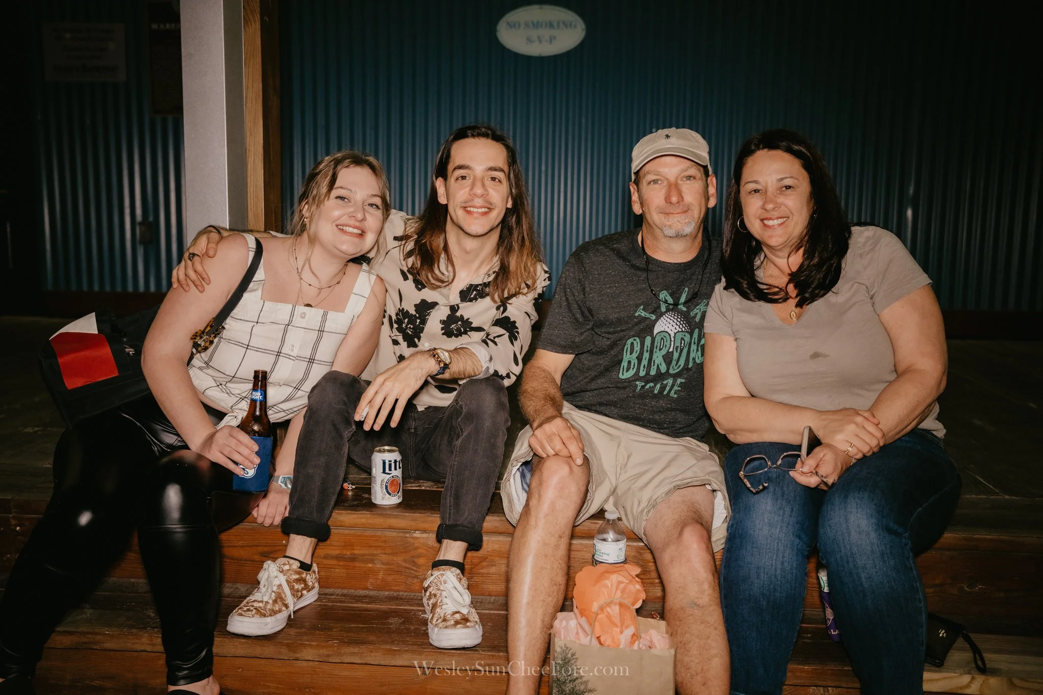 Four people sitting on wooden steps, smiling, holding drinks, with a blue wall and a 'No Smoking' sign behind them.