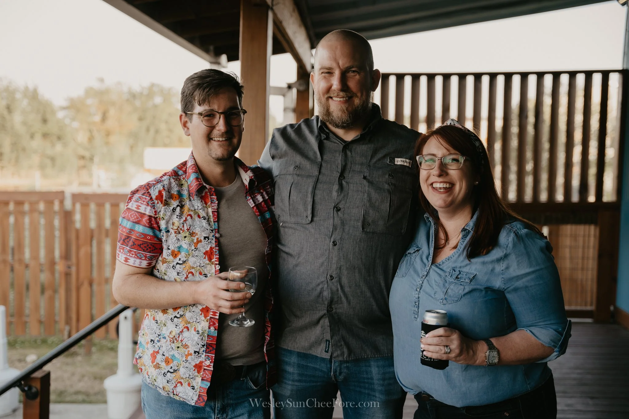 Three people smiling and standing together on a porch, with two men and one woman, each holding a drink, dressed casually.