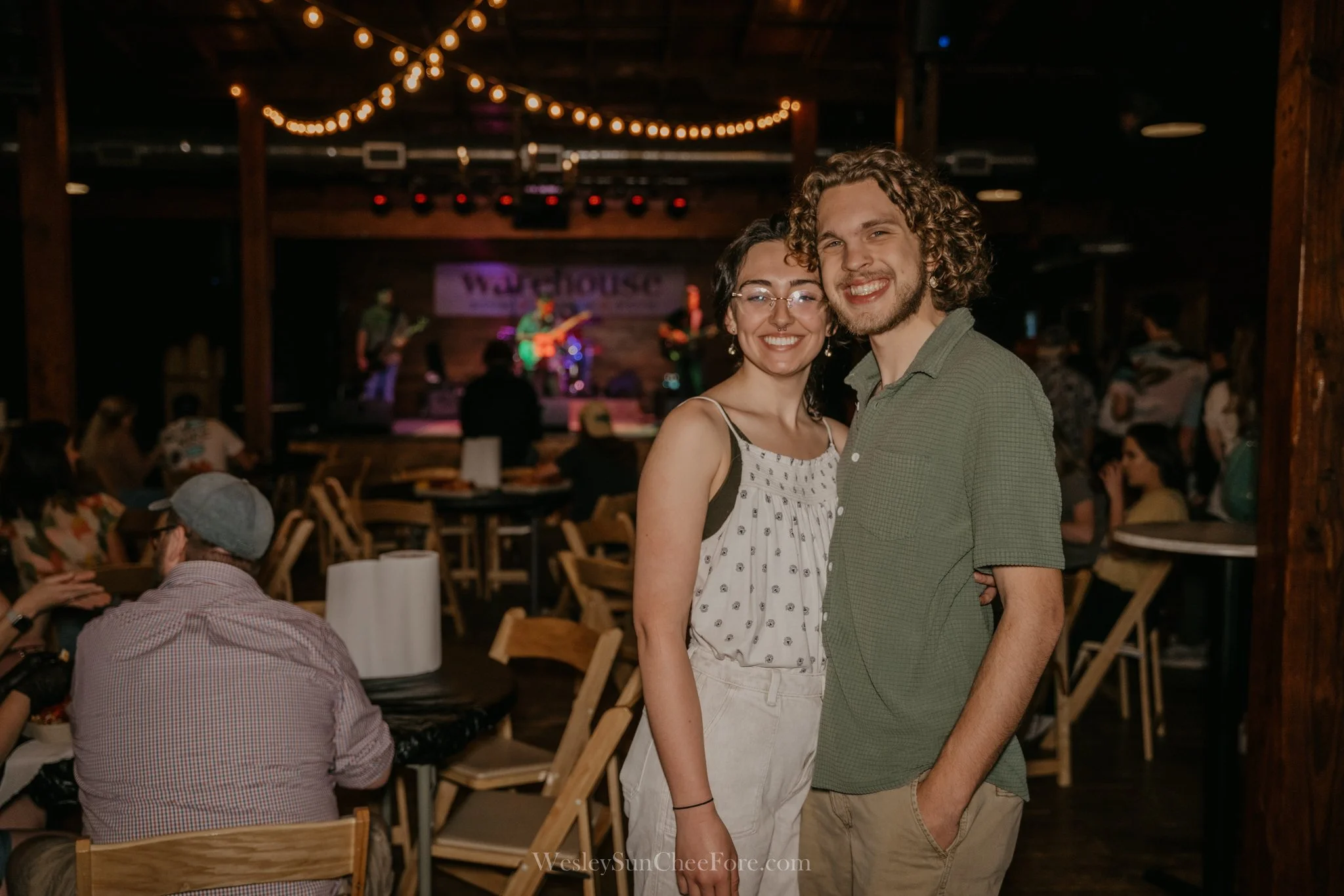 A smiling couple, a woman with glasses and a man with curly hair, pose together at a lively indoor event with a music stage in the background, string lights hanging overhead, and tables filled with people.