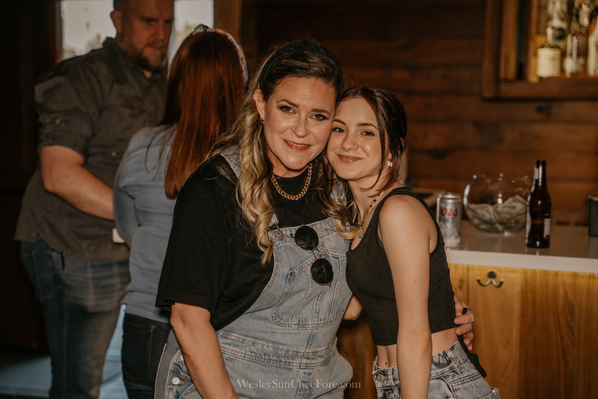 Two women smiling and embracing at a social gathering in a wooden interior space, with two other people in the background.