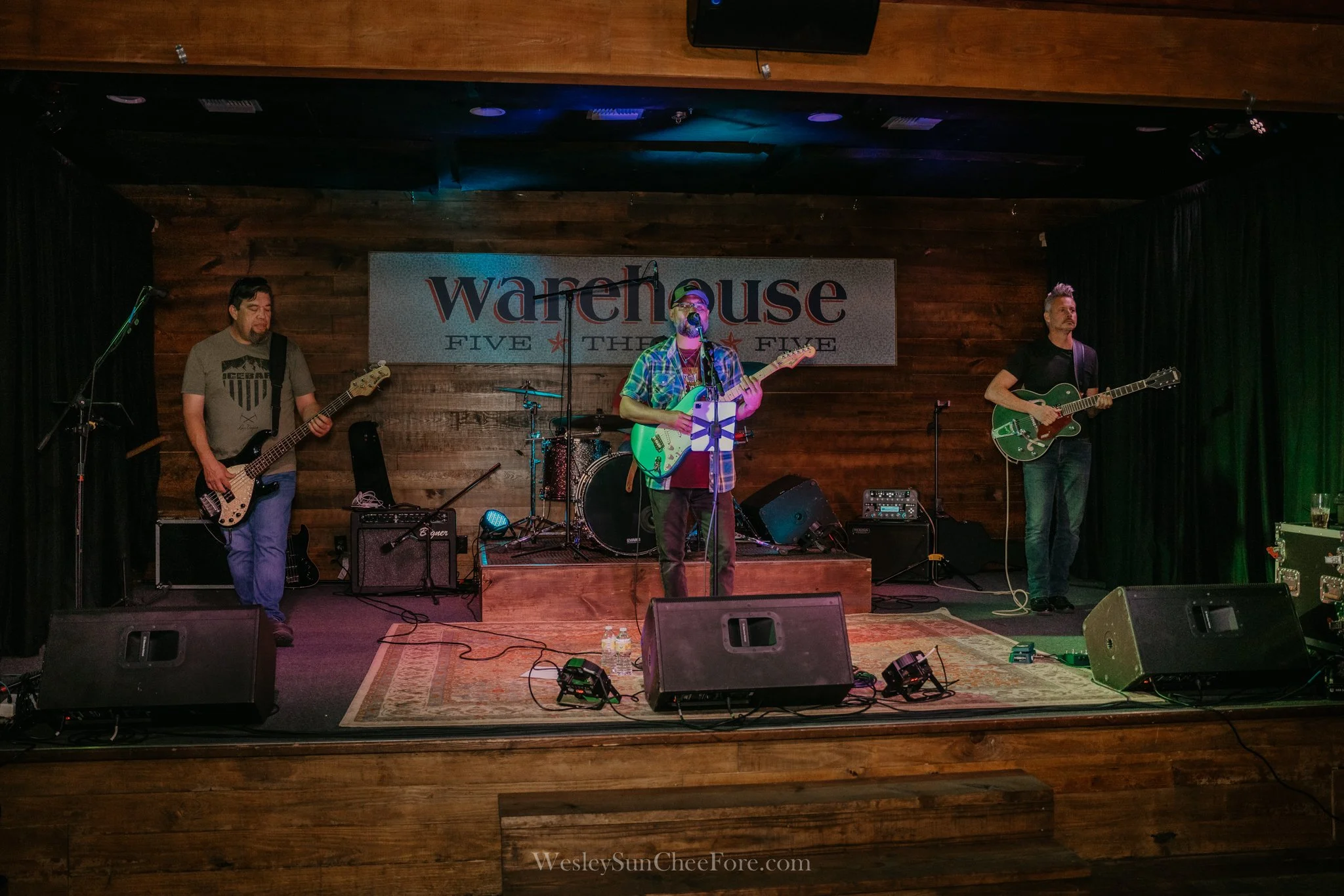 A live band performing on stage at Warehouse Five. Three musicians are playing guitars, with a drum set in the background. The stage has a wooden backdrop and signage that reads "warehouse five."