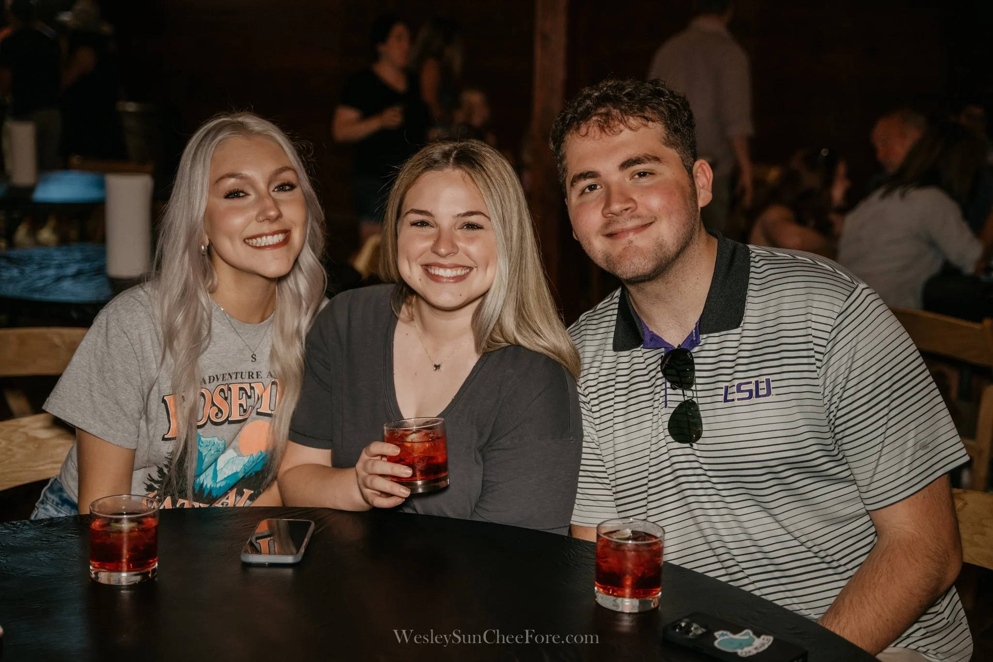 Three young adults smiling at a table with three glasses of red drink, in a dimly lit bar or restaurant setting.