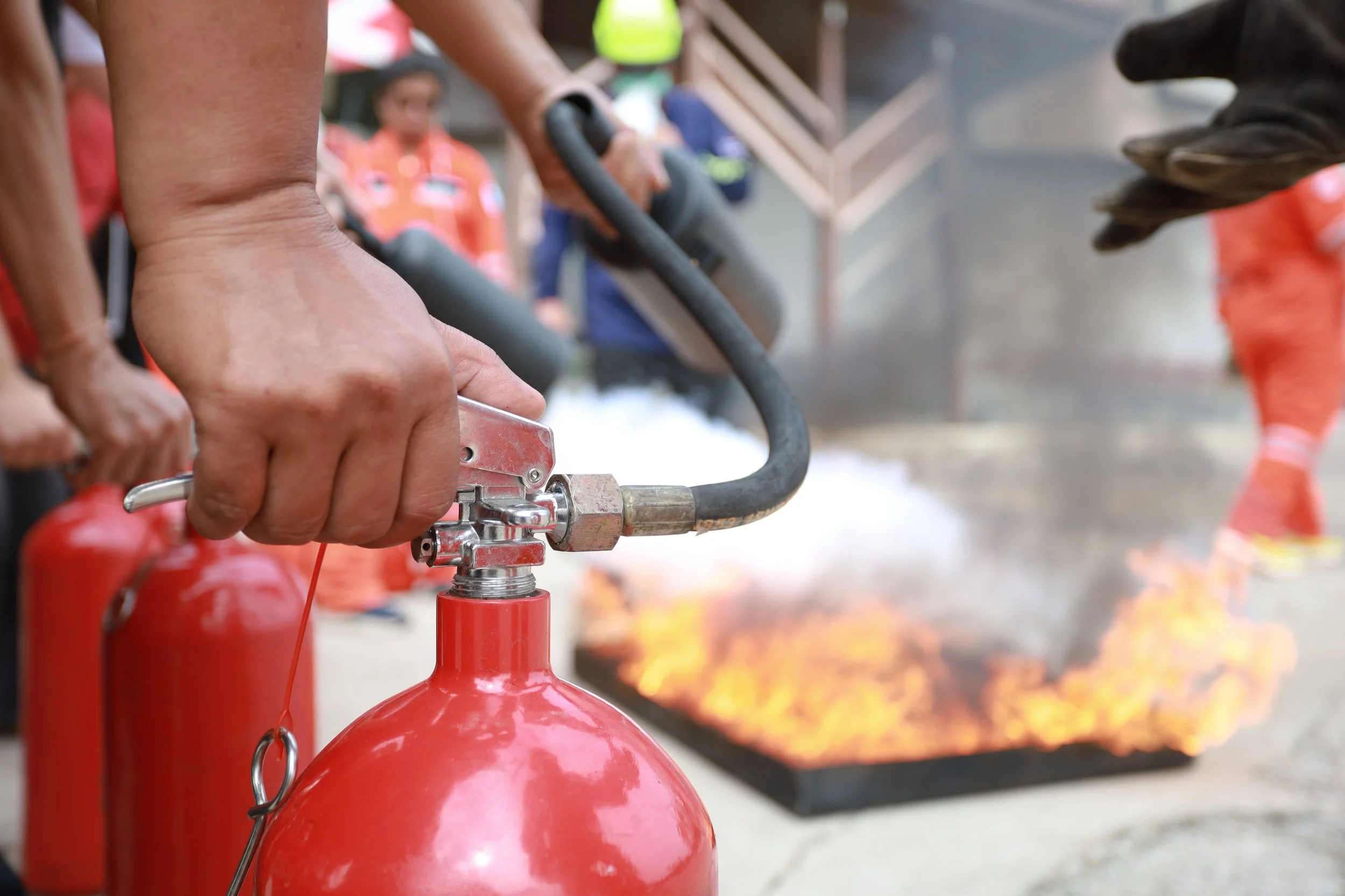 Close-up of a person's hand holding a fire extinguisher nozzle, with a fire burning in the background.