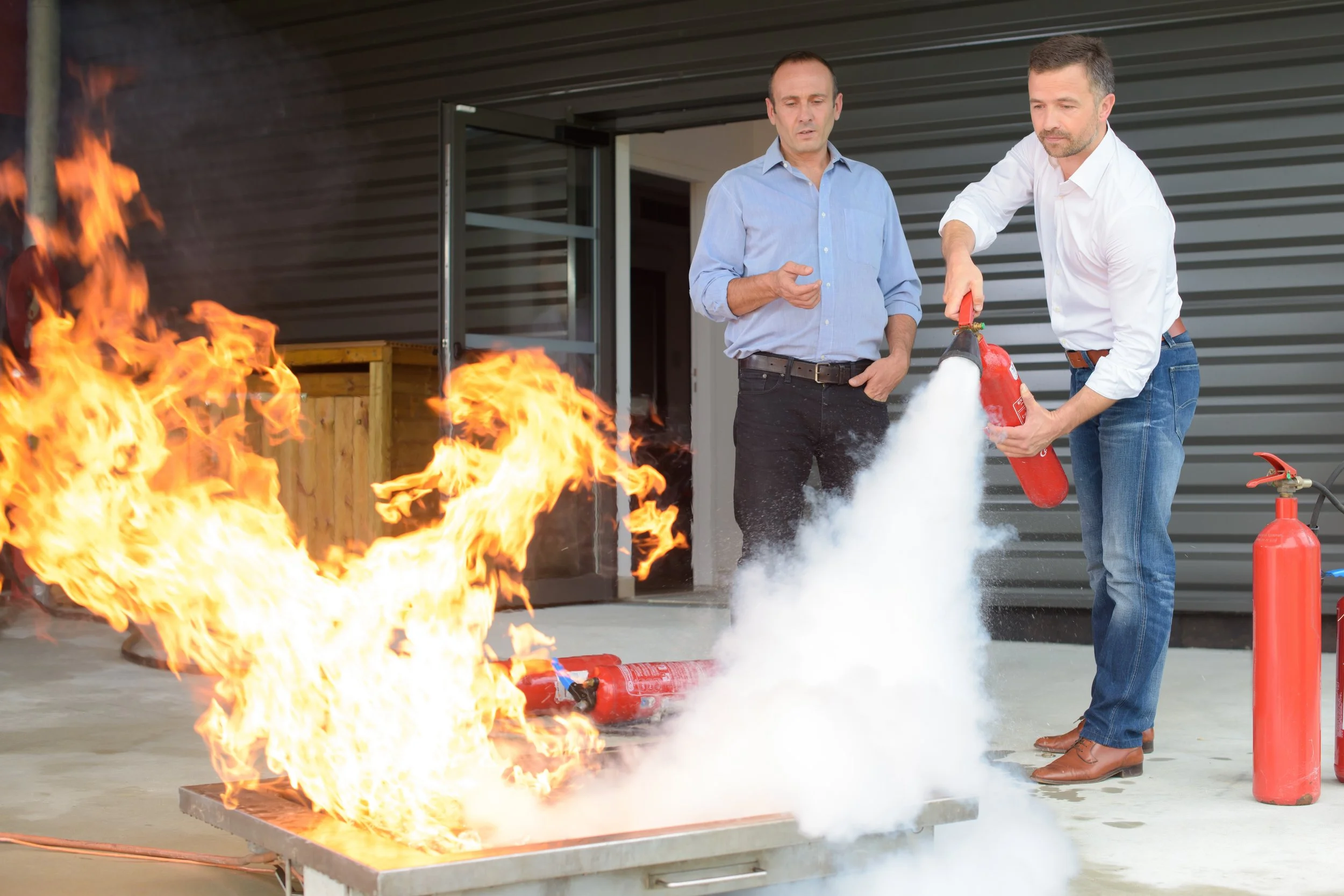 Two men practicing fire safety as one uses a fire extinguisher to put out a small fire on a table outside a building.