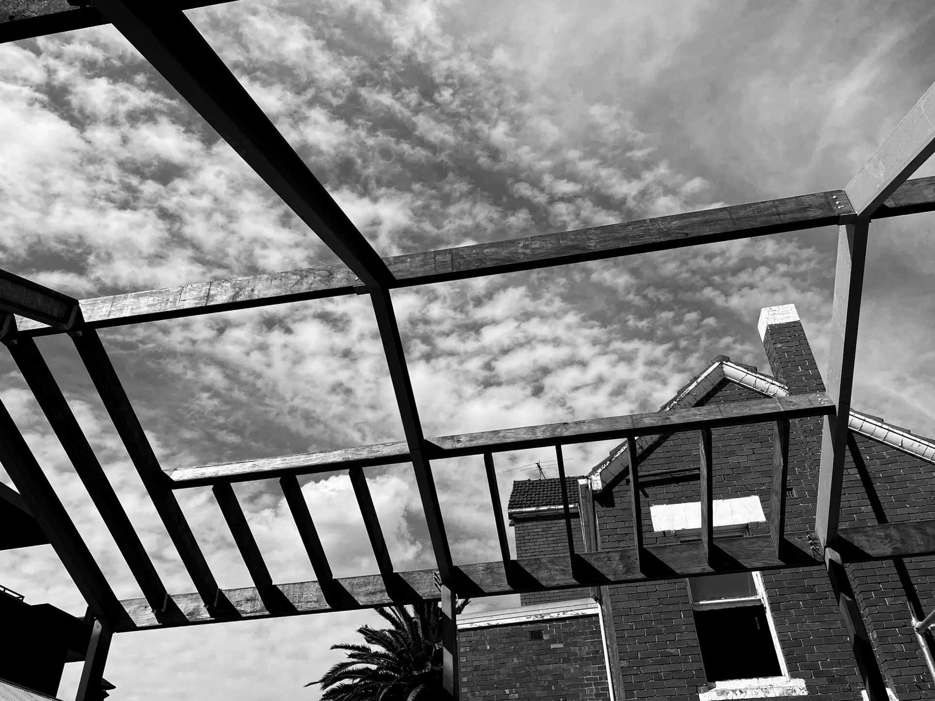 Black and white photo of a house under construction with a wooden framework for a future balcony or roof extension, a chimney, a window, and a partly cloudy sky overhead.