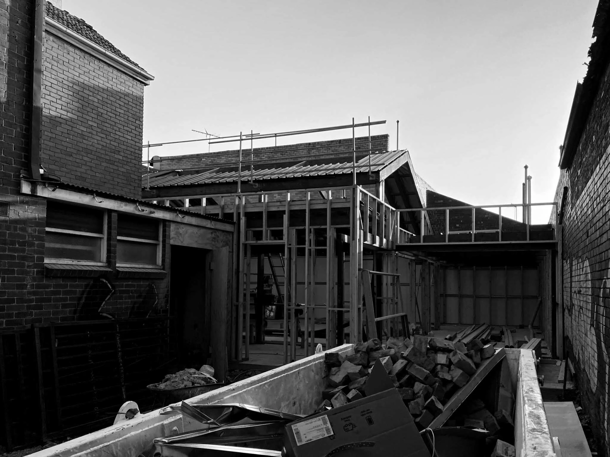 Construction site with wooden framework for building extension, bricks and construction materials scattered around, and adjacent brick buildings.
