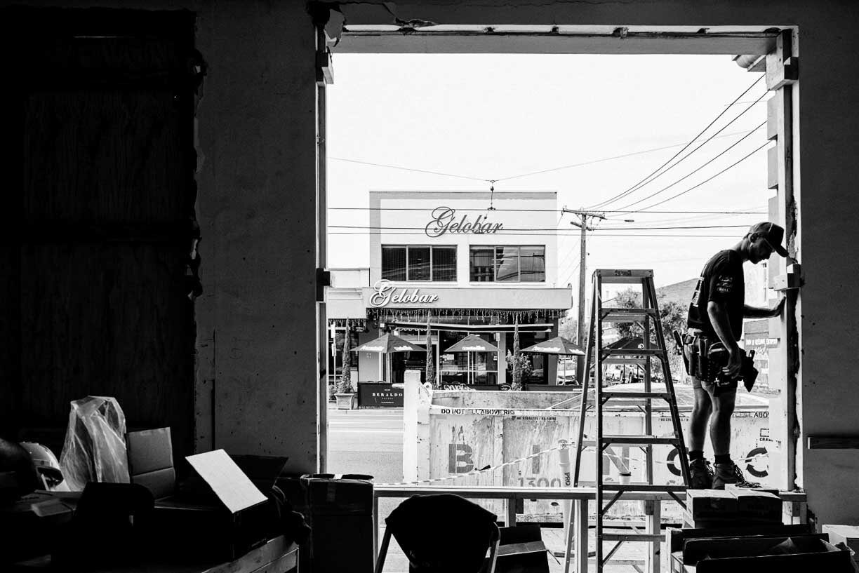A construction worker standing on a ladder inside a building under renovation, looking outside through a large window opening, with a street scene visible beyond including a restaurant called Gelobar.