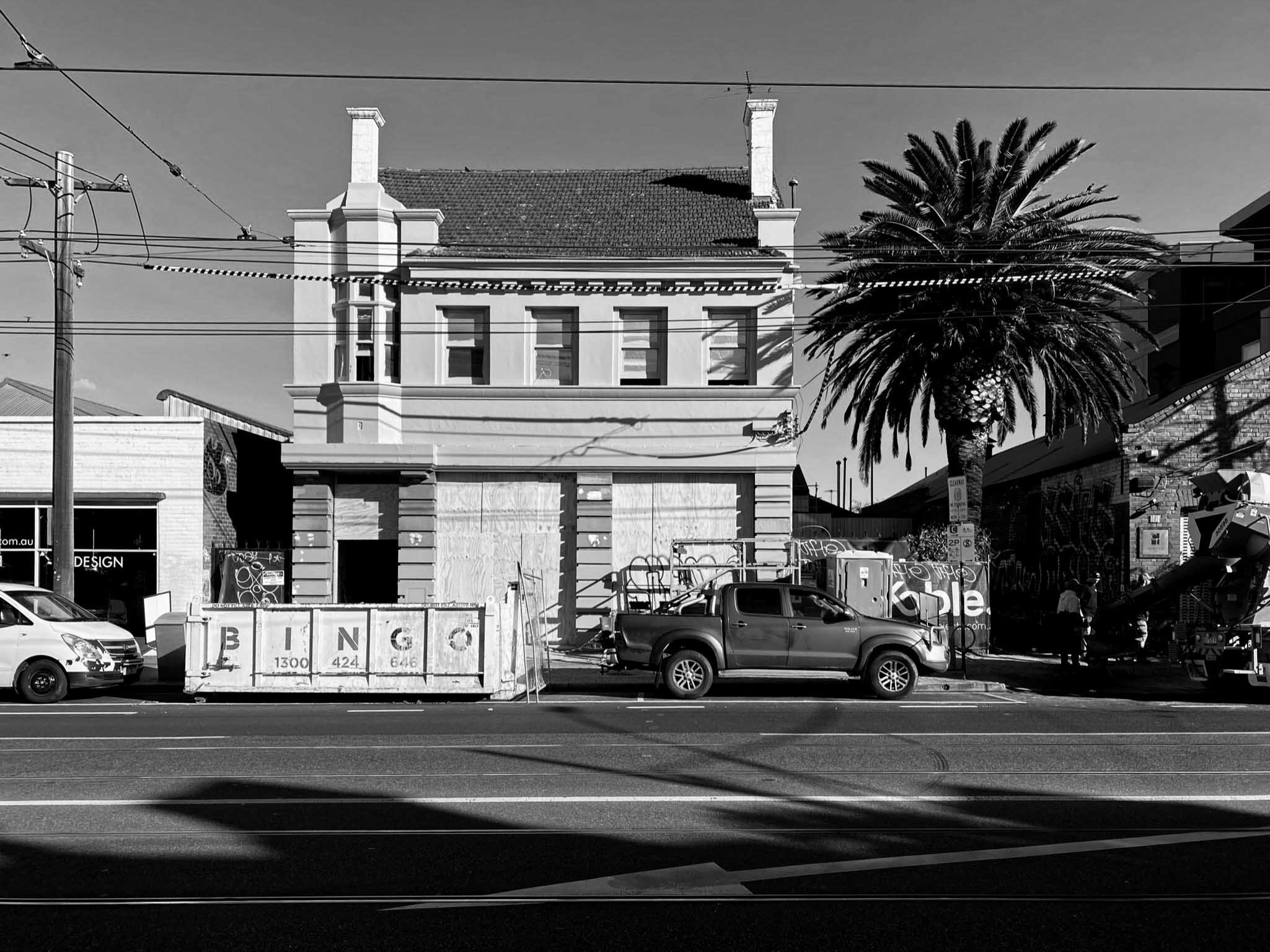 Black and white street scene with a mix of buildings, including a two-story house with boarded-up windows, a palm tree, construction barriers, and parked vehicles.