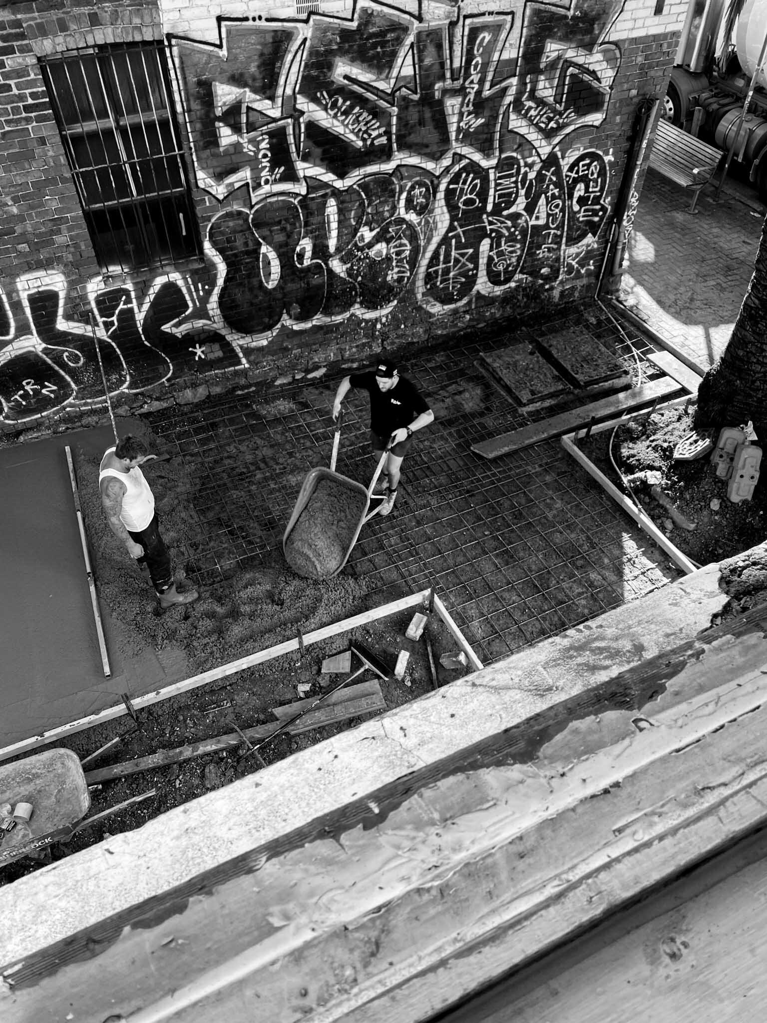 Two construction workers working on a concrete slab with metal rebar, one pouring concrete with a wheelbarrow and another standing nearby. A graffiti-covered brick wall is in the background.