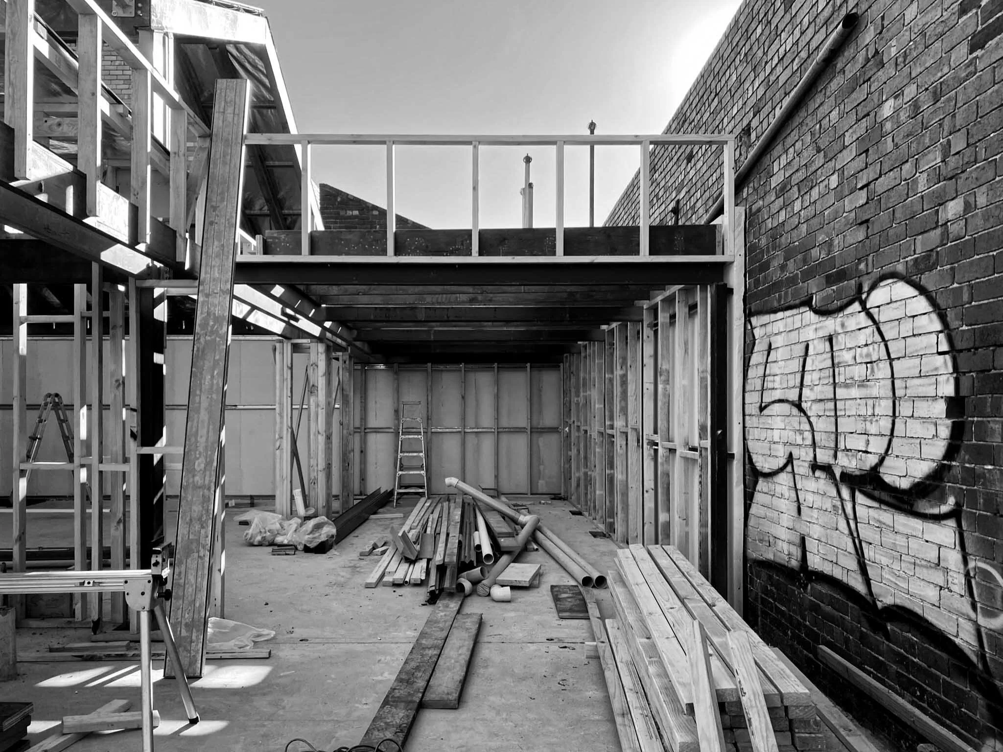 Construction site with wooden framing, metal pipes, ladders, and building materials, next to a graffiti-covered brick wall.