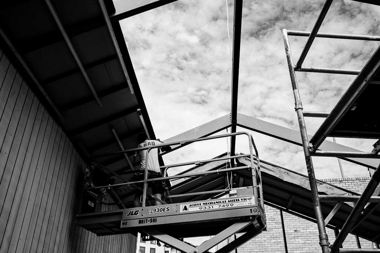 A worker in a safety harness standing on a cherry picker lift, working on a house roof under construction, with scaffolding nearby and a partly cloudy sky above.