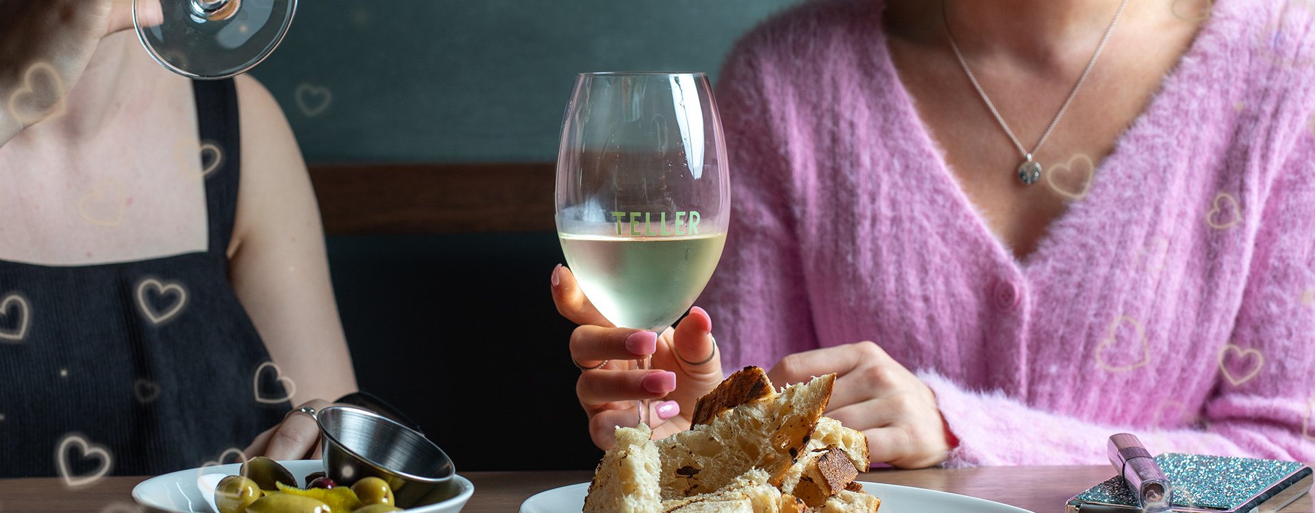 Two women at a table with a glass of white wine, bread, olives, and a glittery pink purse.