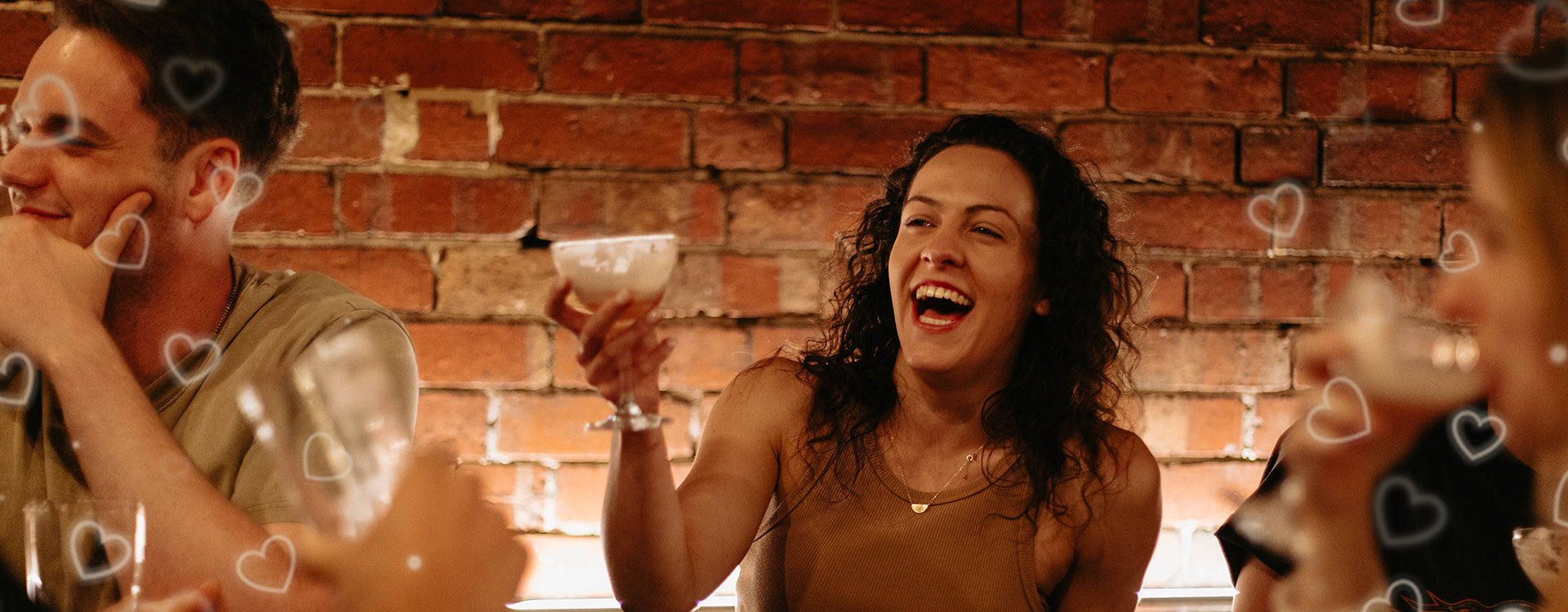 A woman laughing and holding a cocktail at a social gathering with a brick wall background.