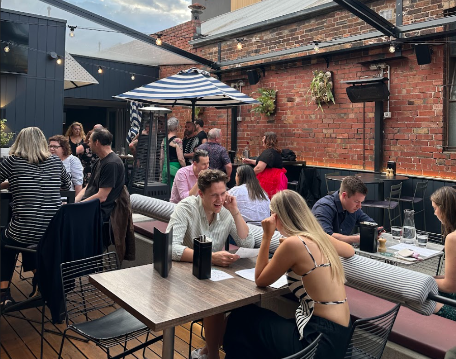 People dining and socializing at an outdoor restaurant patio with string lights, a striped umbrella, and brick walls.