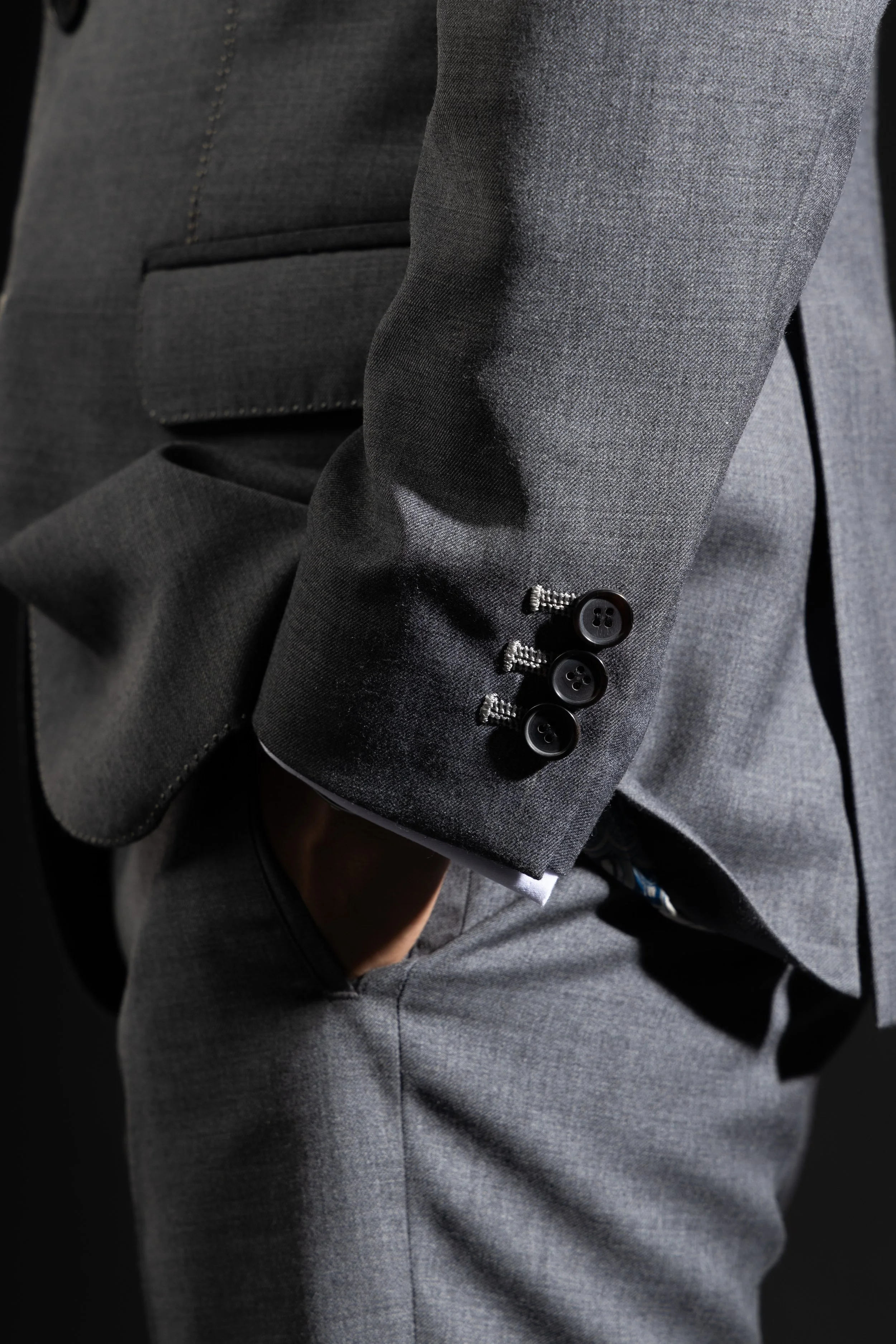 A side close up of the button holes on a grey suit jacket's sleeve, focusing on intricate hand finishing