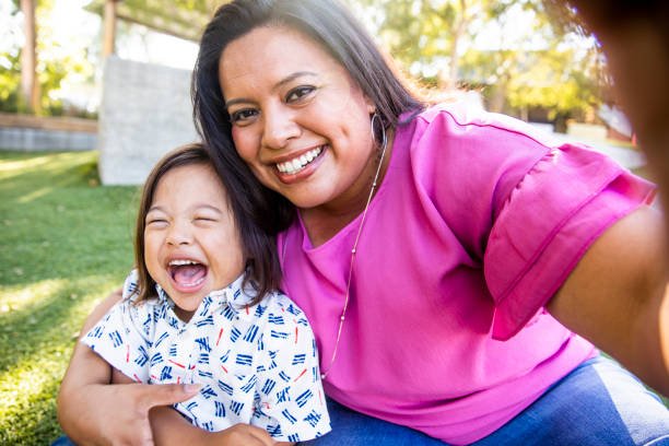 A woman and a young girl taking a selfie outdoors in a park, both smiling and showing happiness.