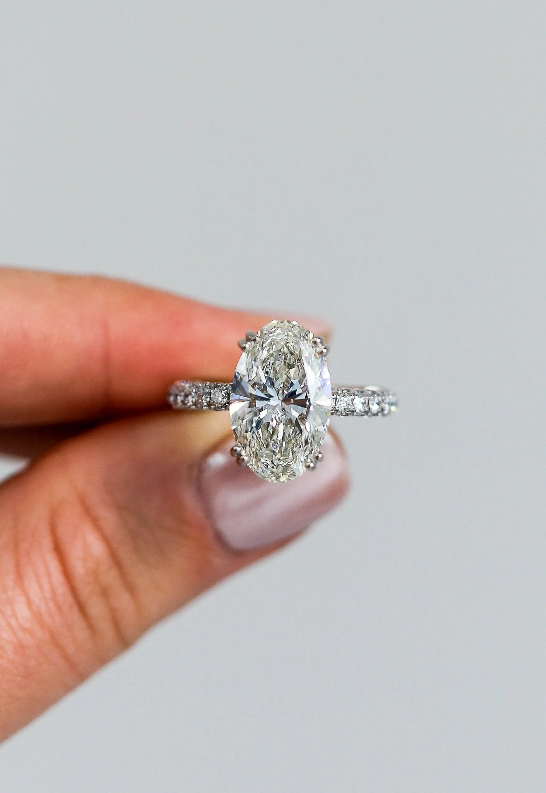 Close-up of a person's hand holding an oval-shaped diamond ring with smaller diamonds on the band against a neutral background.
