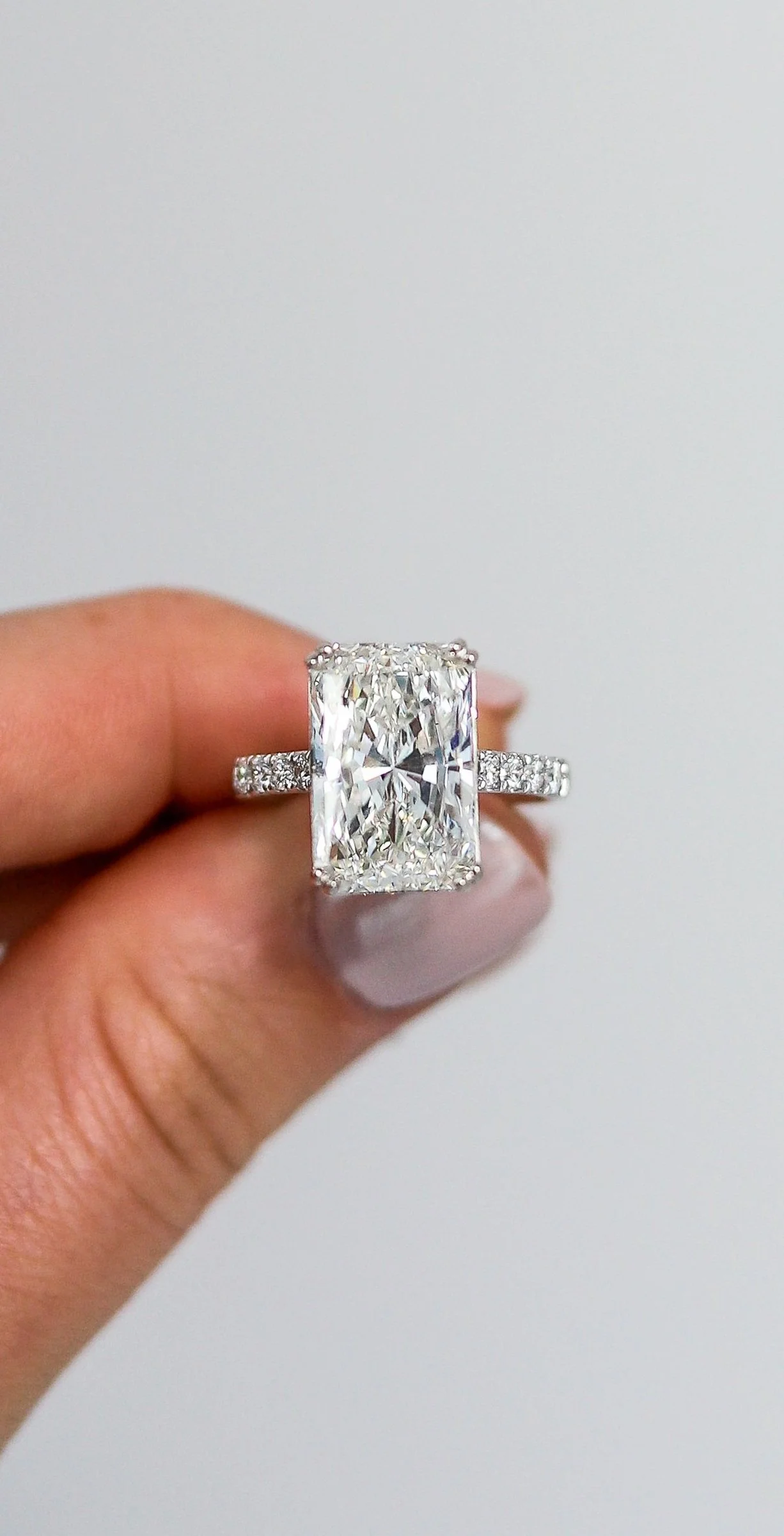 A hand holding a large rectangular cut diamond engagement ring with a diamond-studded shank, against a plain white background.