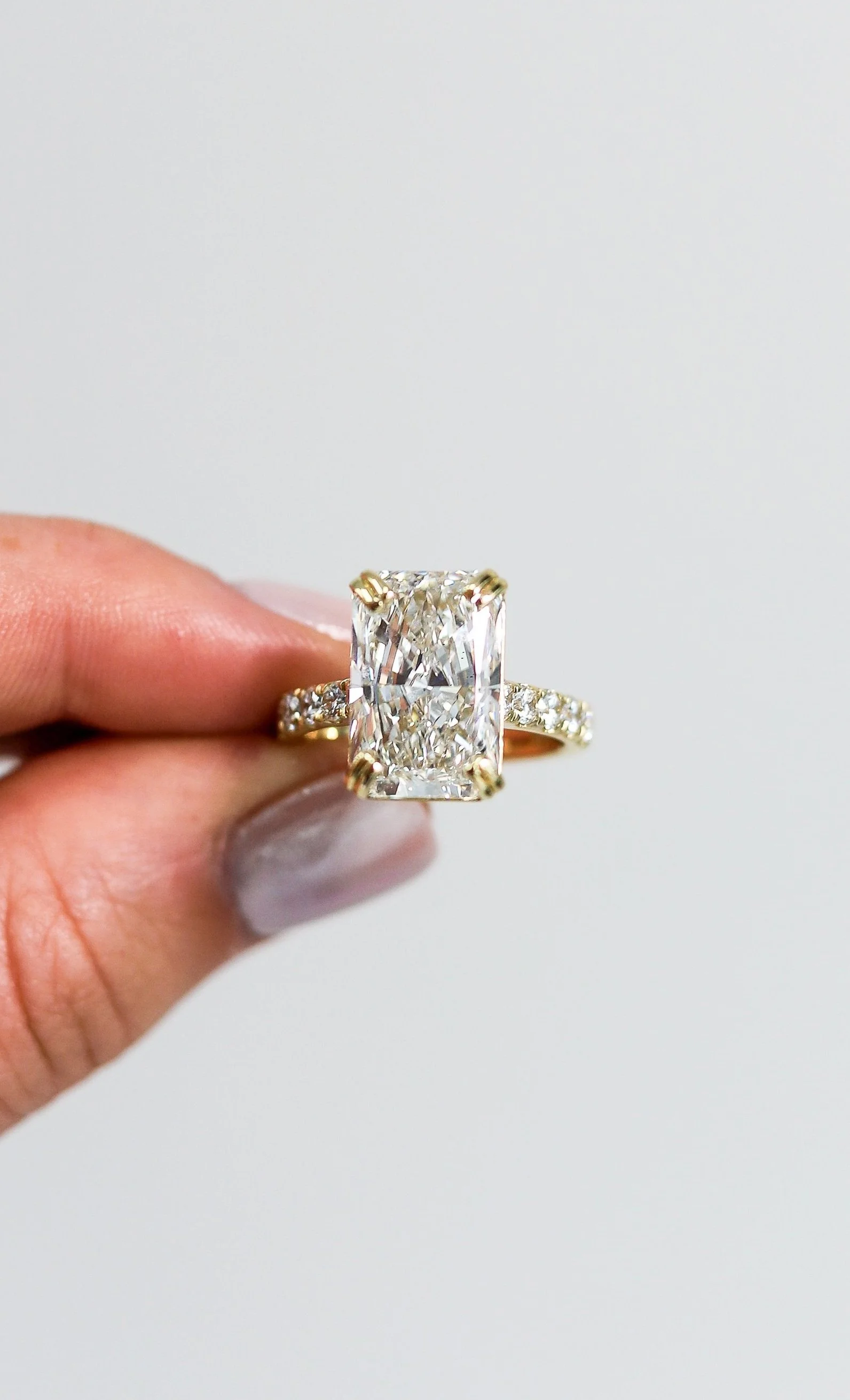 Close-up of a hand holding a gold ring with a large rectangular-cut diamond and smaller diamonds on the band, with a plain white background.