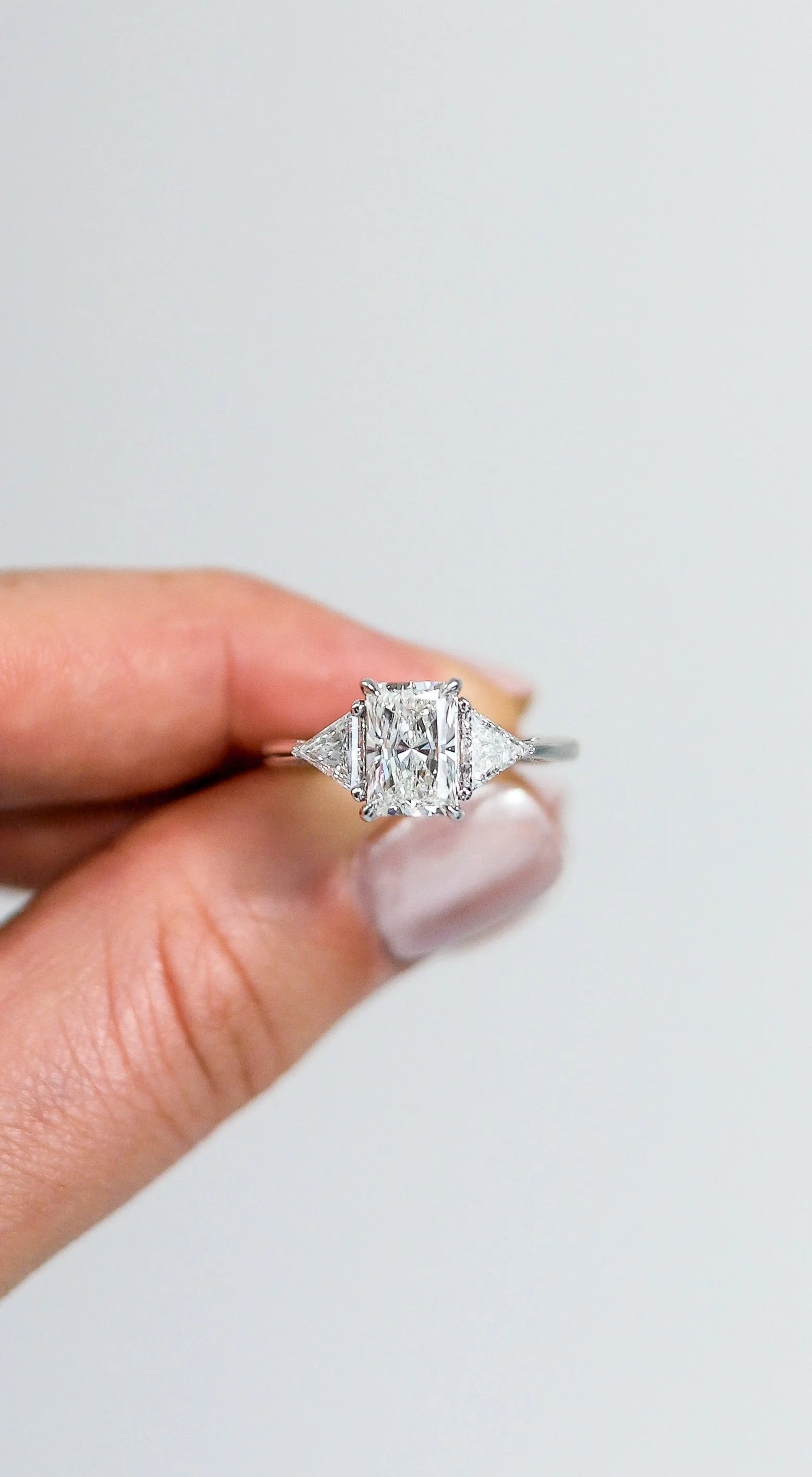 Close-up of a diamond engagement ring with an emerald-cut center stone and two triangular side stones, held between fingers.