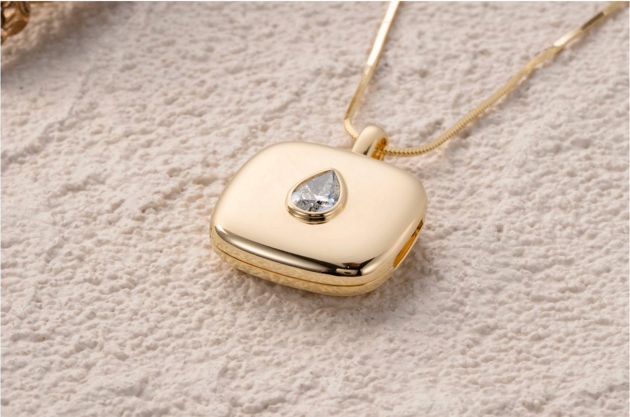 Everdear Erinnerungsdiamant-Medaillon aus 14 Karat Gelbgold mit birnenförmigem Kremationsdiamanten, gezeigt auf einer weich strukturierten Oberfläche mit feiner goldener Schlangenkette