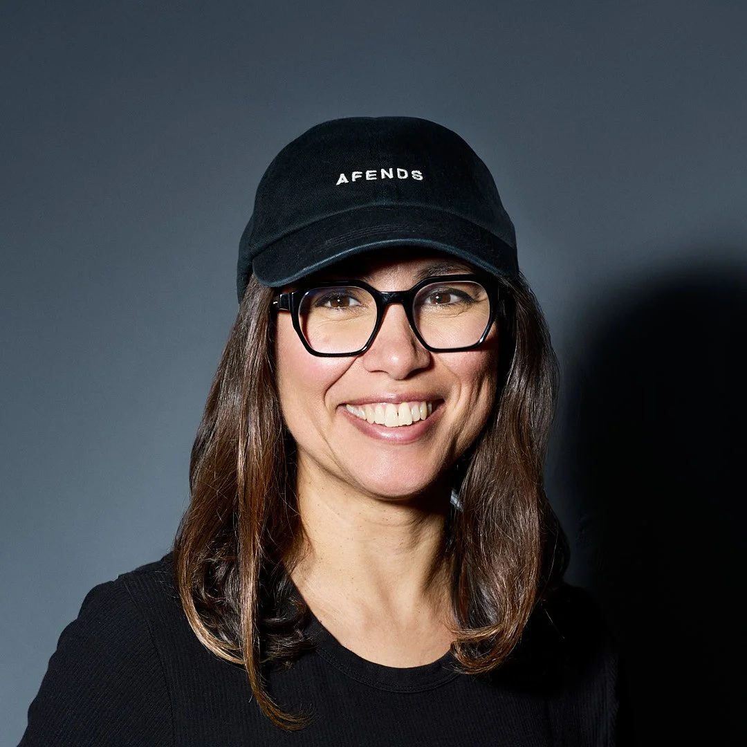 Claudia Martinez, photographer,  woman wearing a black cap with 'AFENDS' written on it, black glasses, and a black shirt, smiling against a dark gray background.