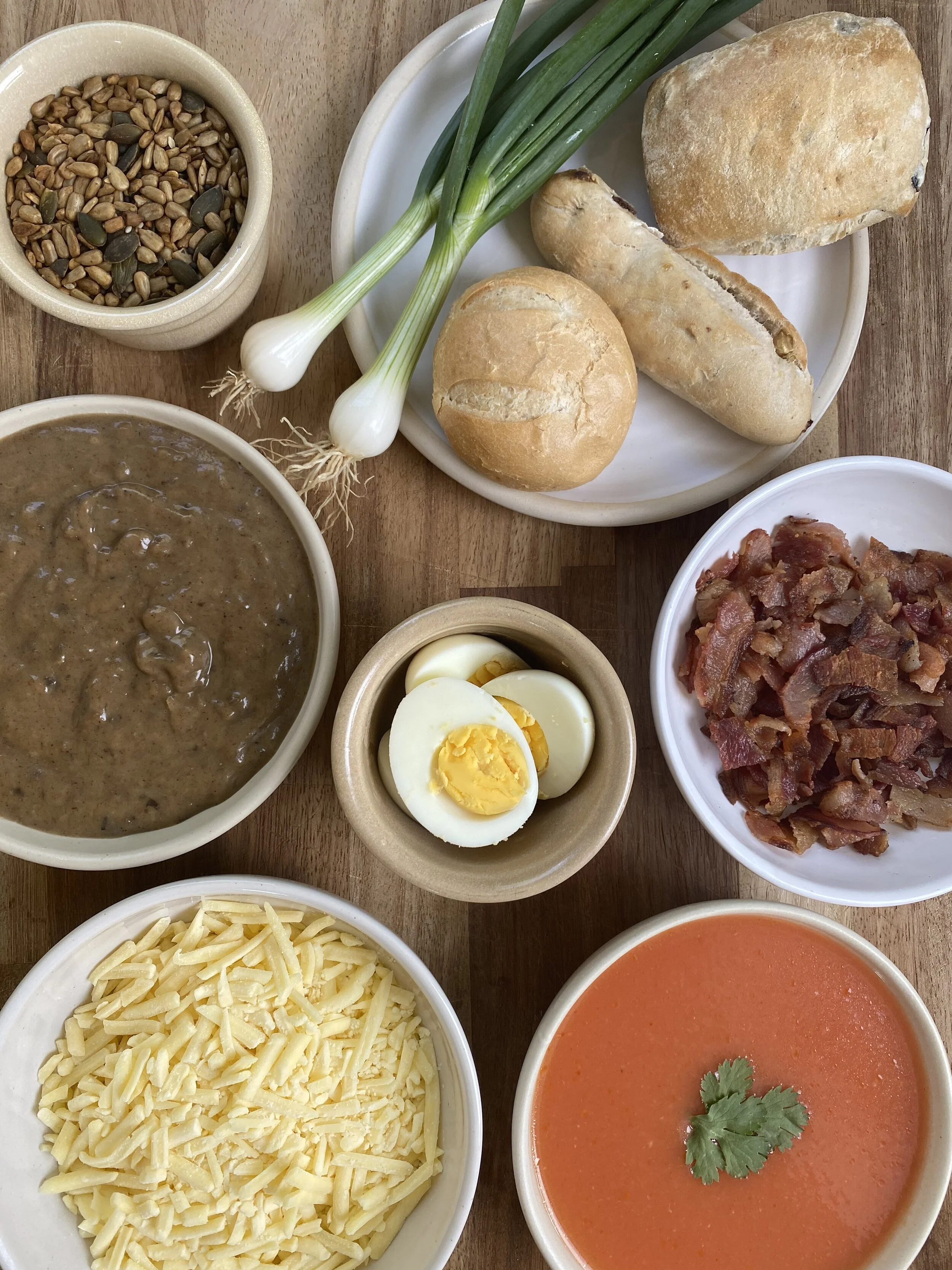 Bowls of shredded cheese, bacon bits, Tomato soup with cilantro with boiled egg, a Wild mushroom soup with bread rolls, and toppings.