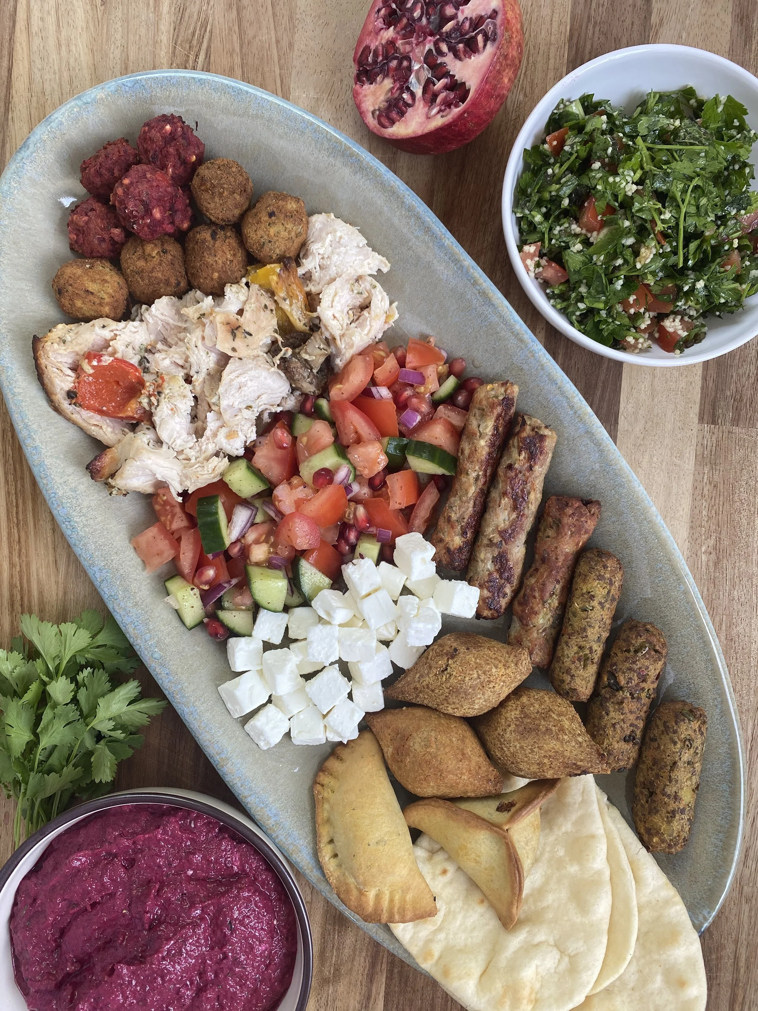 A large serving platter of Middle Eastern nibbles with falafel, grilled chicken, salad, feta cheese, pita bread, and grape leaves, accompanied by a bowl of Beetroot & Za'atar dip and a bowl of Tabbouleh salad, with pomegranate seeds.