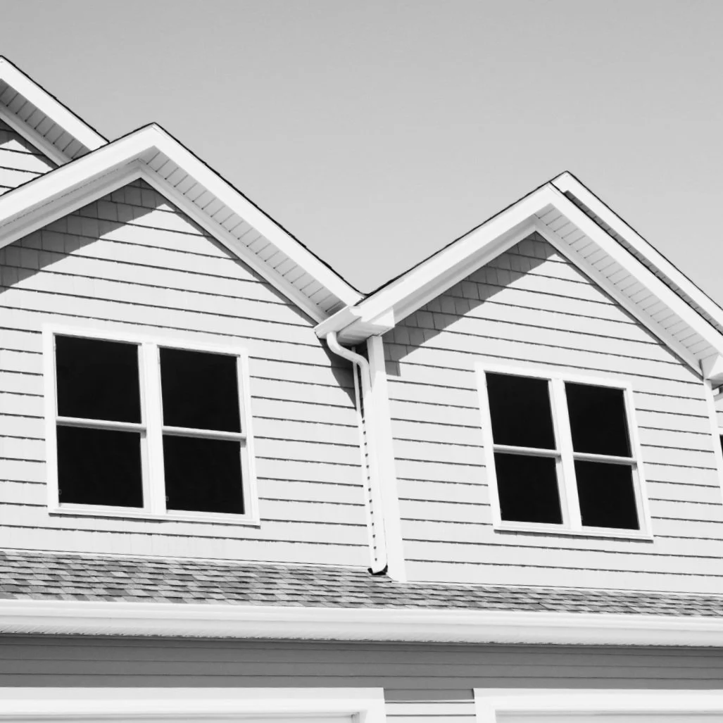 Black and white photo of the upper part of a house with siding, two double-pane windows, and a roof with shingles.