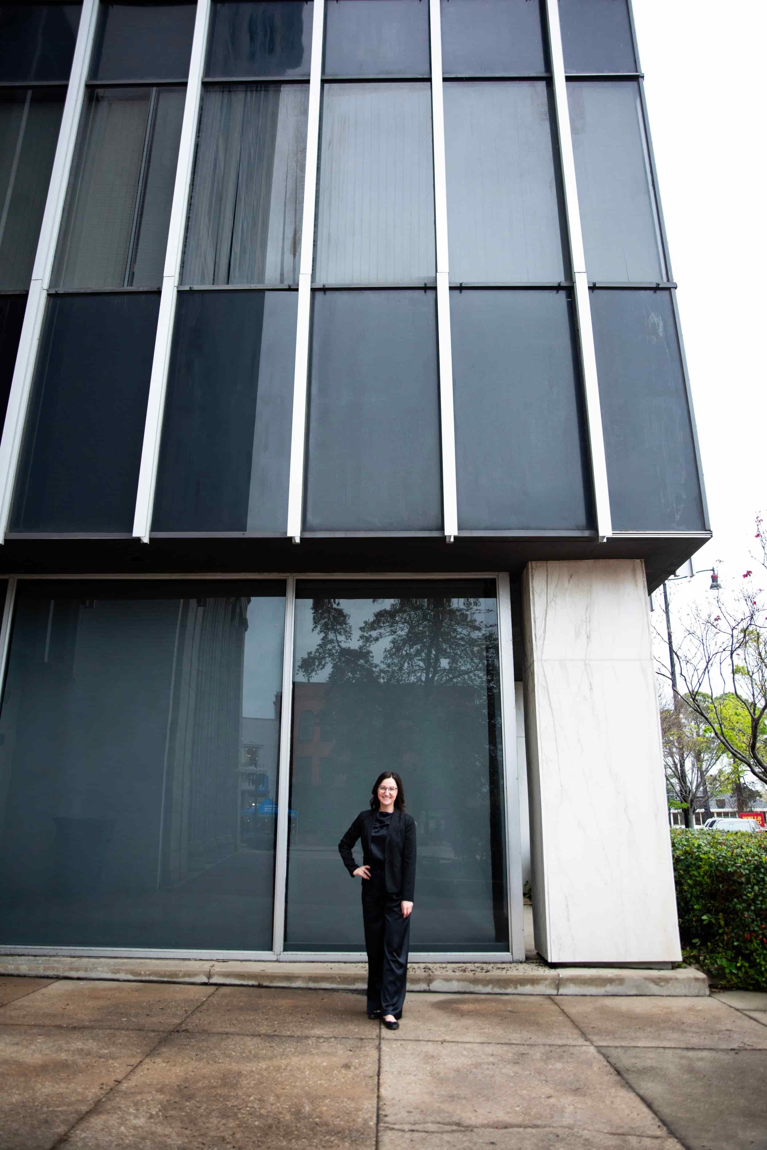 Woman in a black suit standing outside a modern building with large glass windows.