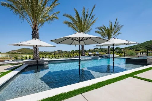 Luxurious outdoor pool with large umbrellas and palm trees under a clear blue sky.