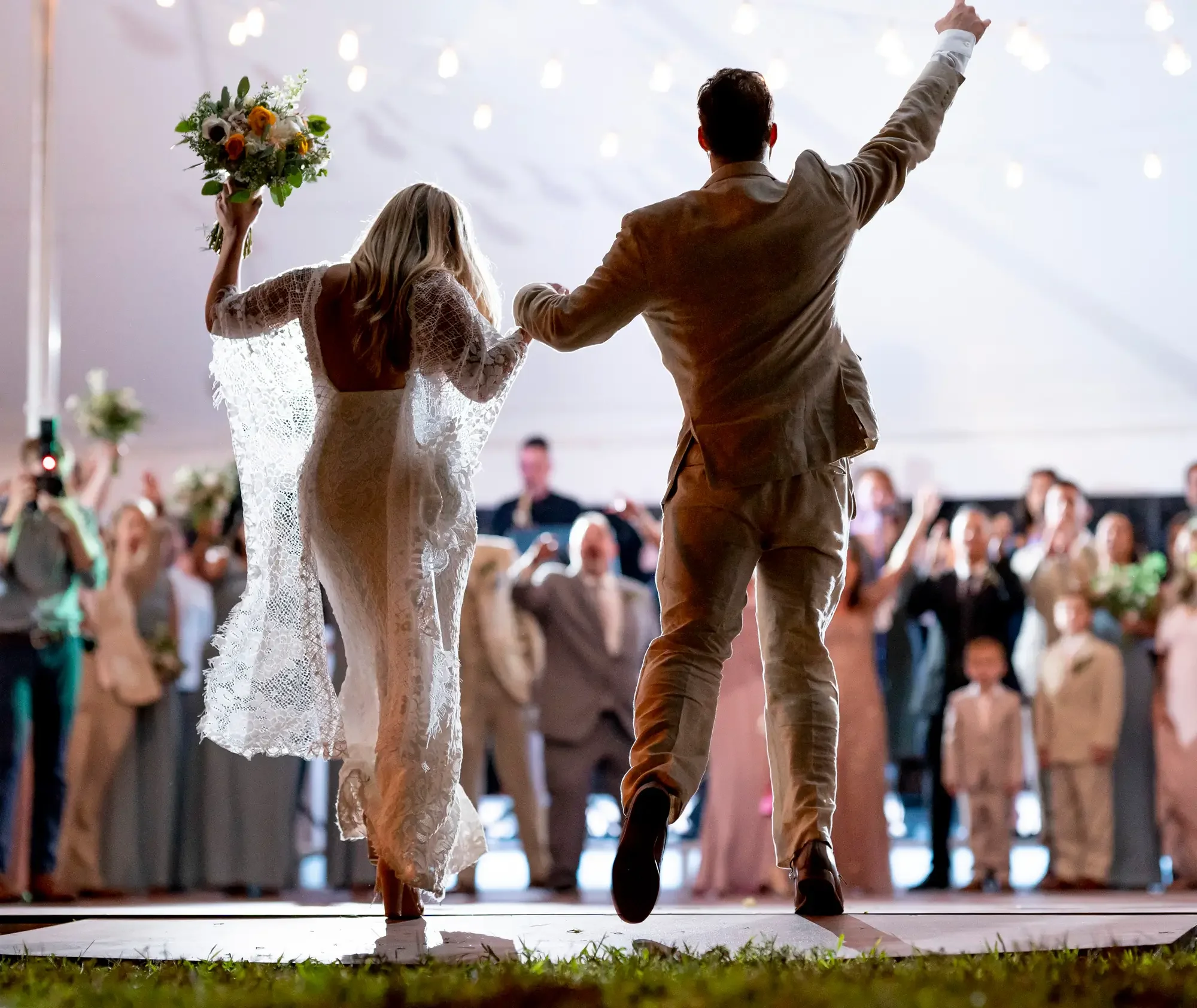 A bride and groom dance together at their wedding reception, with guests watching and taking photos in the background.