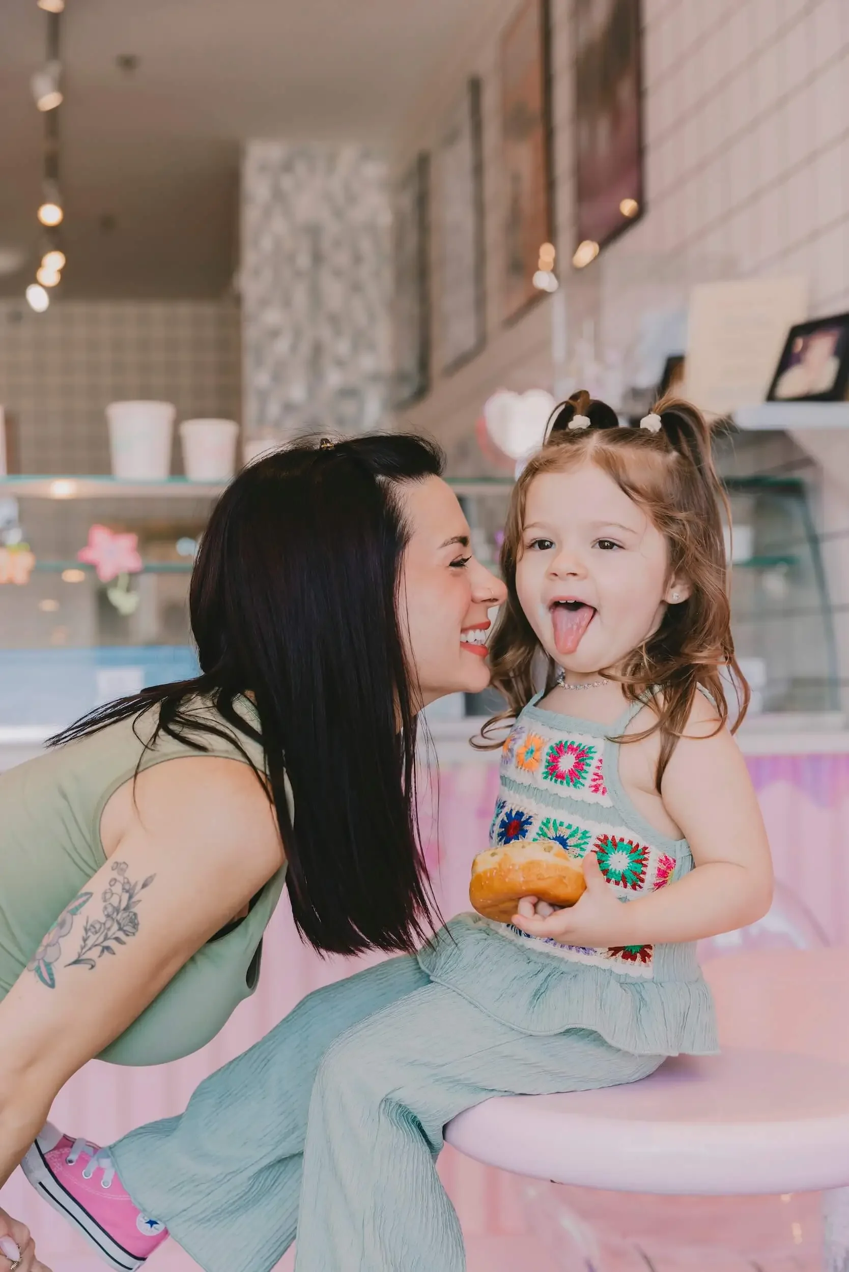 A woman leaning close to a young girl with brown hair in pigtails at an ice cream shop. The girl is sticking her tongue out and holding a donut. The woman has a tattoo on her arm and is smiling.