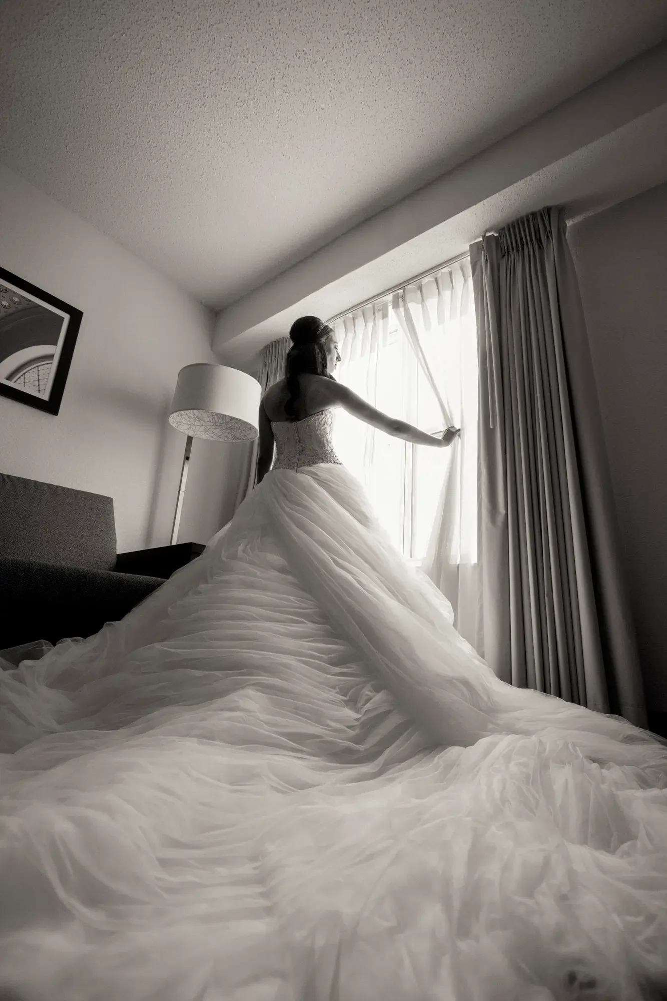 A bride in a strapless wedding dress standing by a window with curtains, looking outside, in a hotel room with a coiled train on the floor.