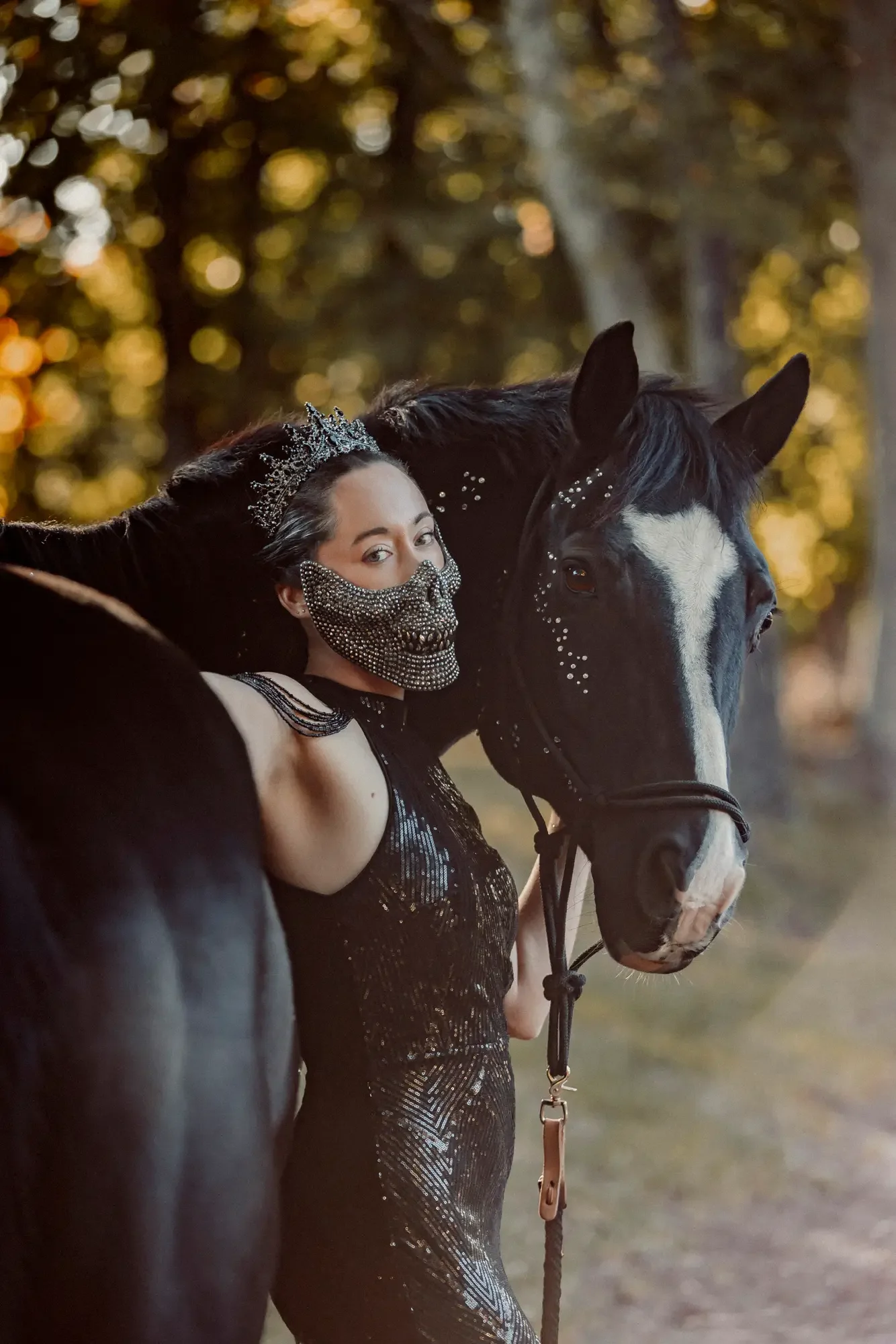 A woman dressed in black with a rhinestone skull face mask and wearing a crown stands next to a black and white horse with white face markings and decorative rhinestones on its face, outdoors during sunset.