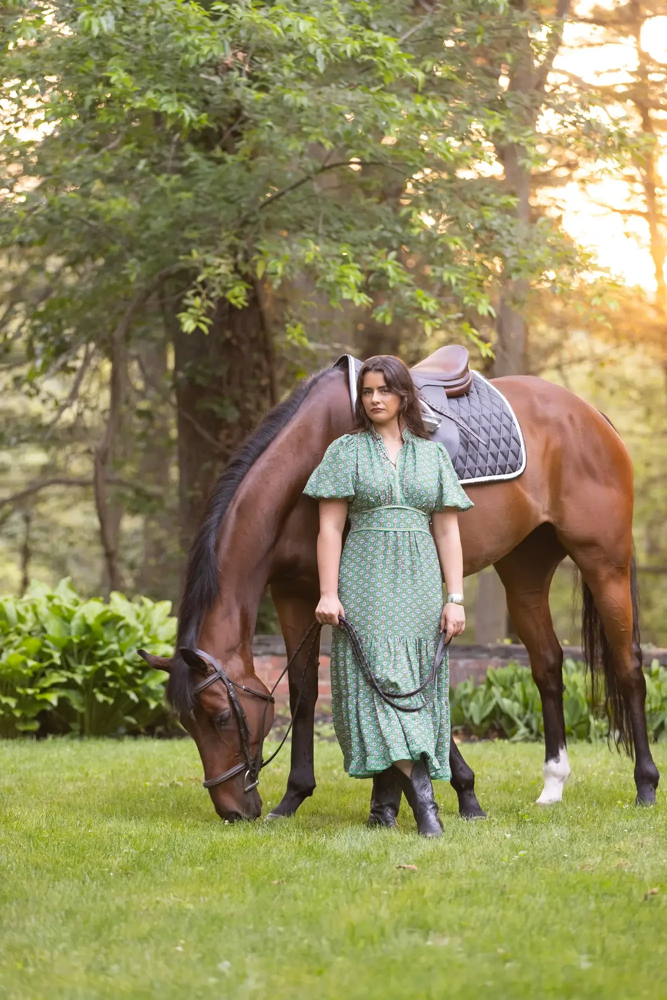 A woman in a green dress holding horse reins, standing next to a chestnut horse with a saddle in a wooded area during sunset.