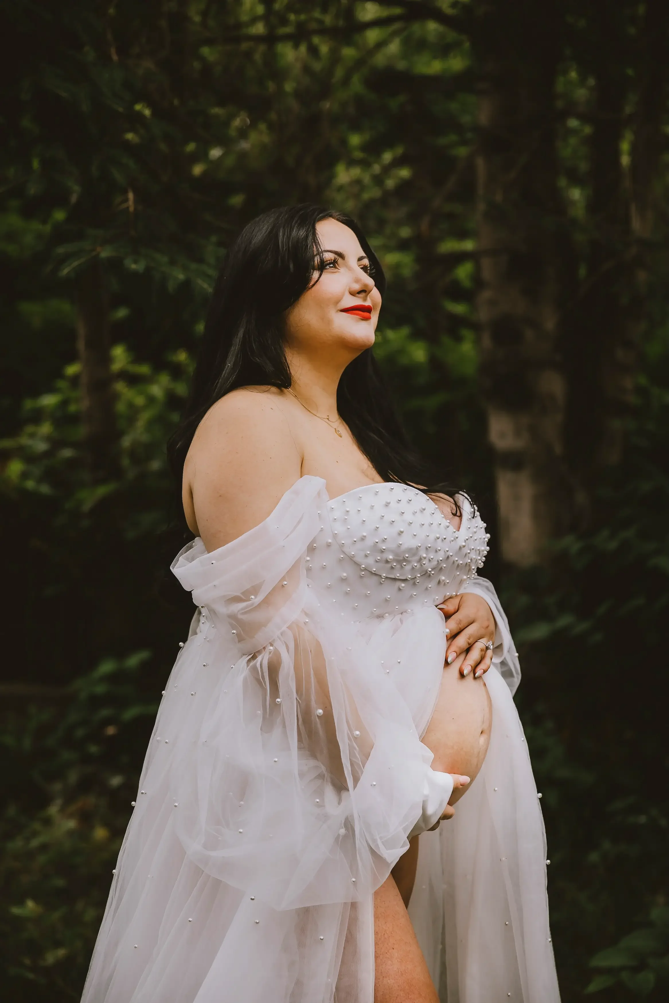 A pregnant woman in a white, pearl-embellished dress with sheer, flowing sleeves, standing outdoors in a forest, gently holding her belly and smiling.