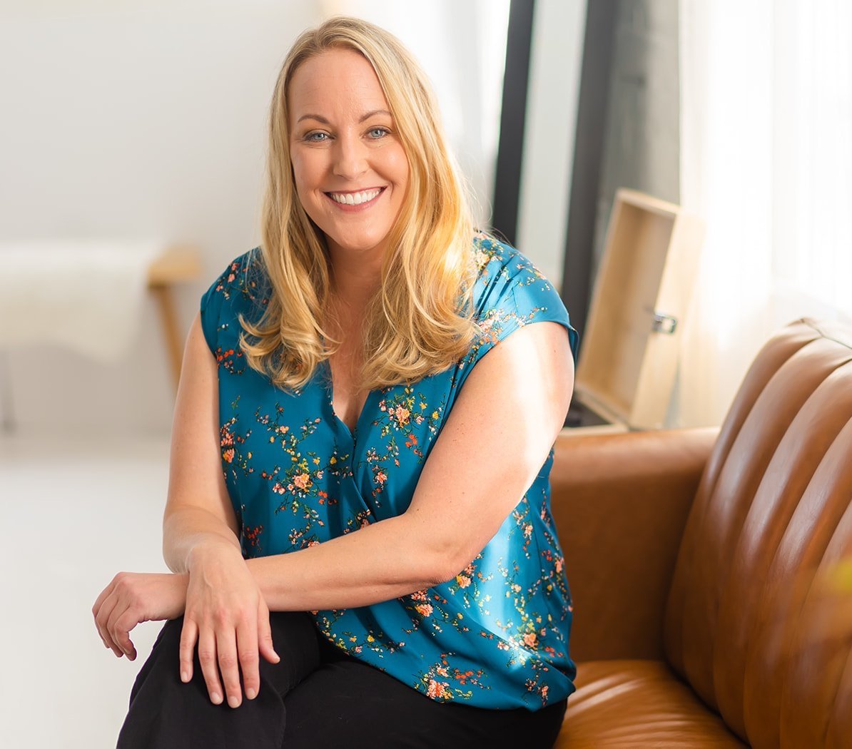 A smiling woman with long blonde hair wearing a blue floral top, sitting on a brown leather couch in a bright room.