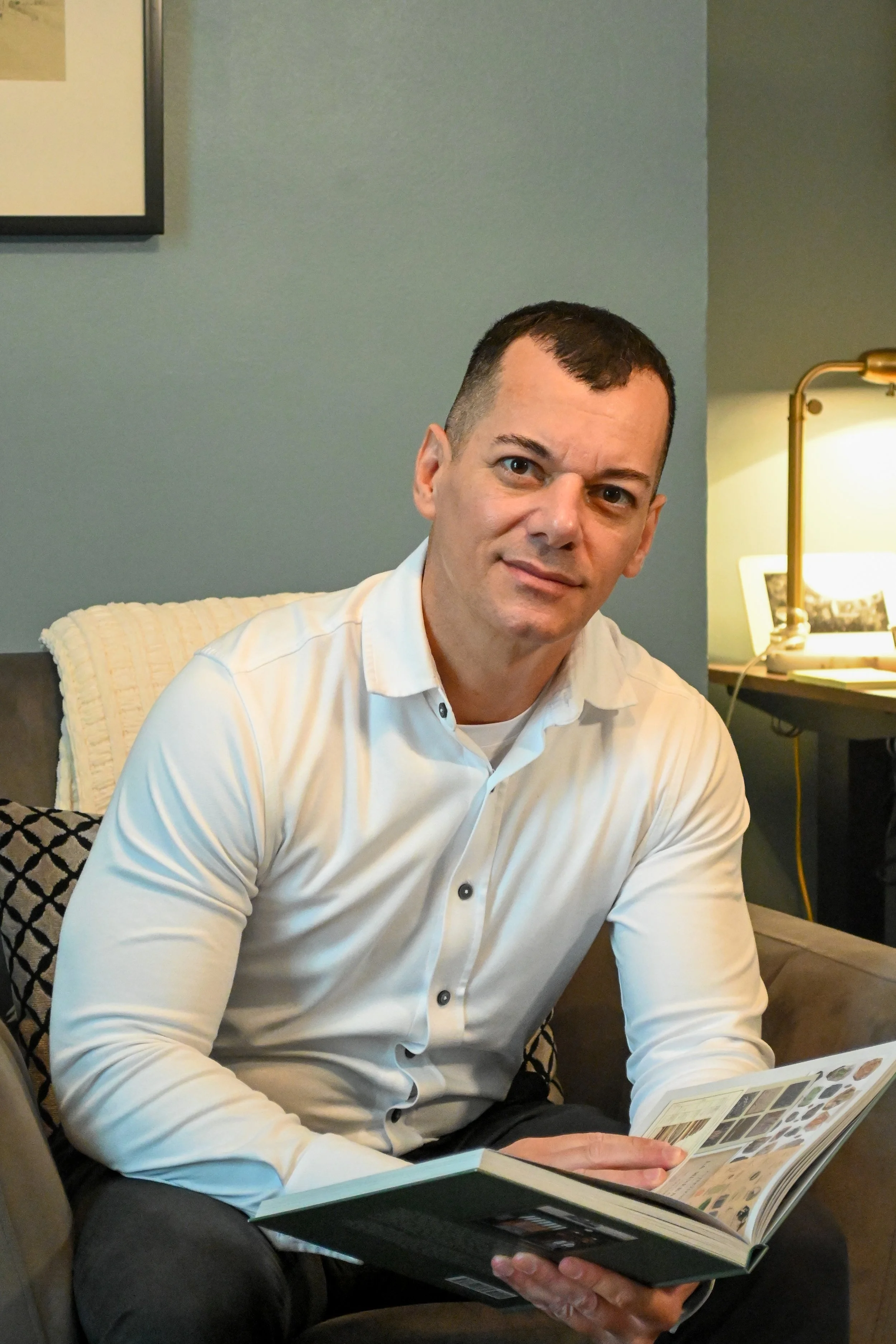 A man in a white shirt sitting on a couch, reading a magazine, with a study lamp and framed picture in the background.