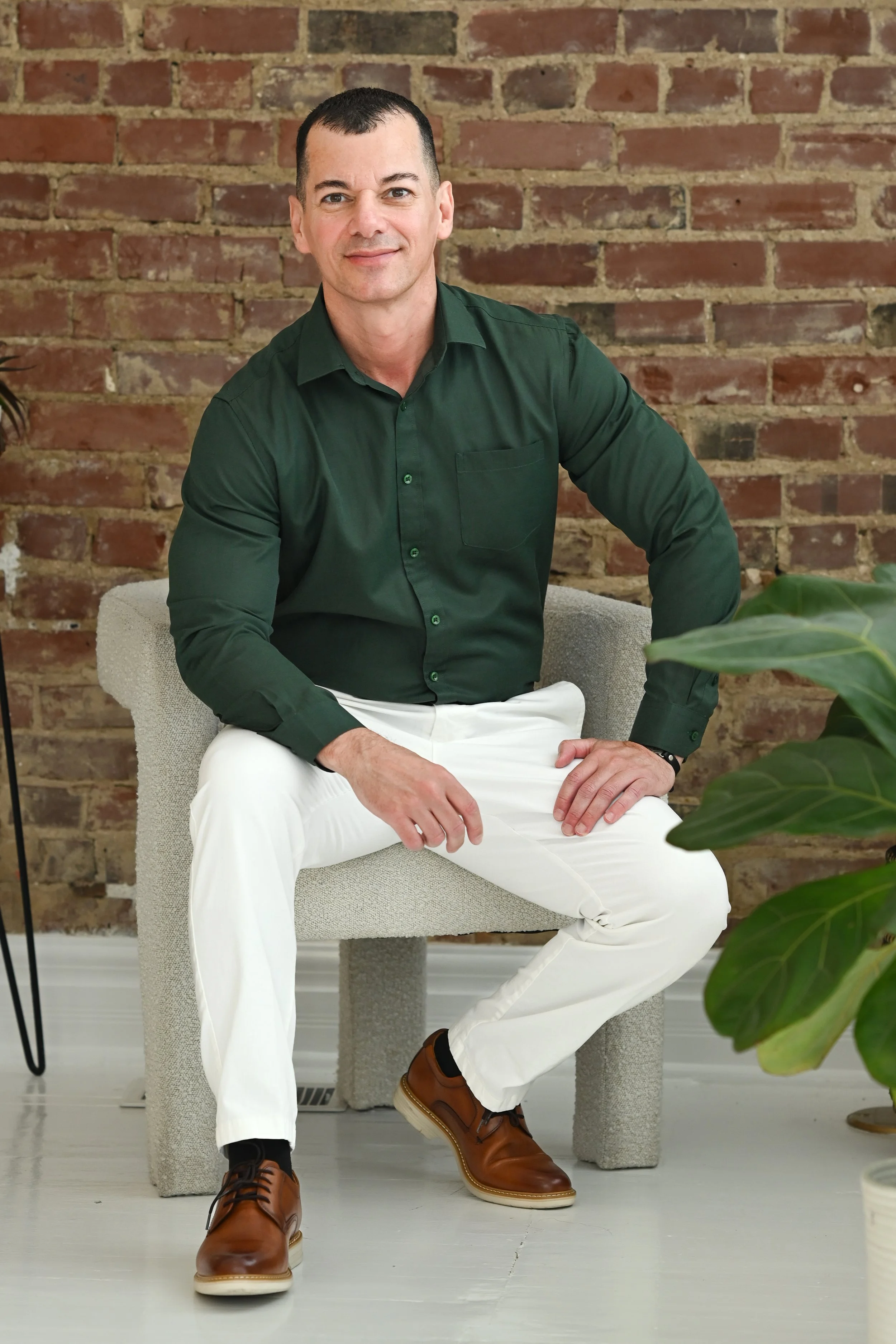 Man with short dark hair wearing a dark green shirt and white pants, sitting on a beige chair in front of a brick wall with some green plants visible.