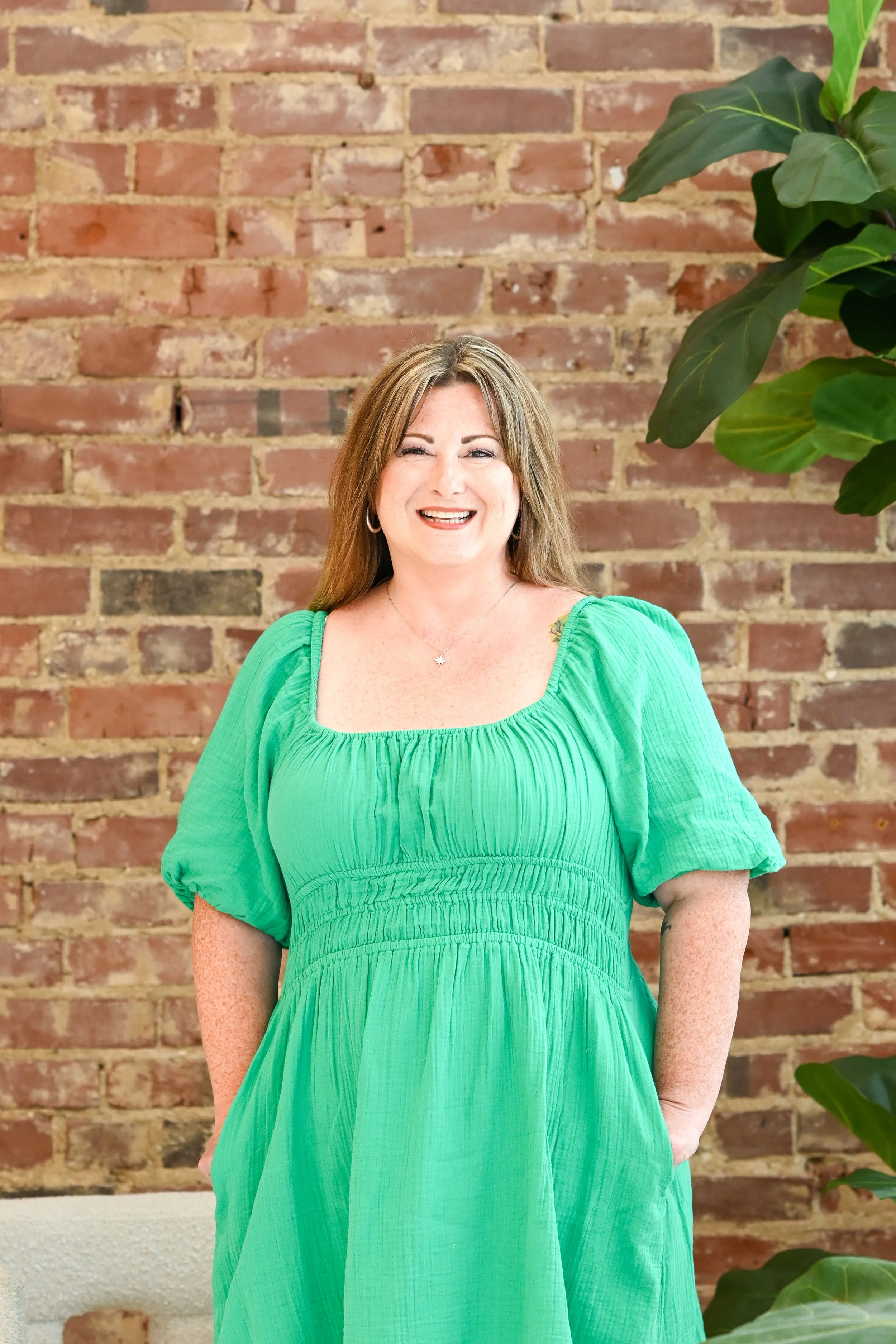 A woman with long, light brown hair and freckles, smiling and wearing a bright green dress, standing in front of a brick wall with a large green plant on the right side.