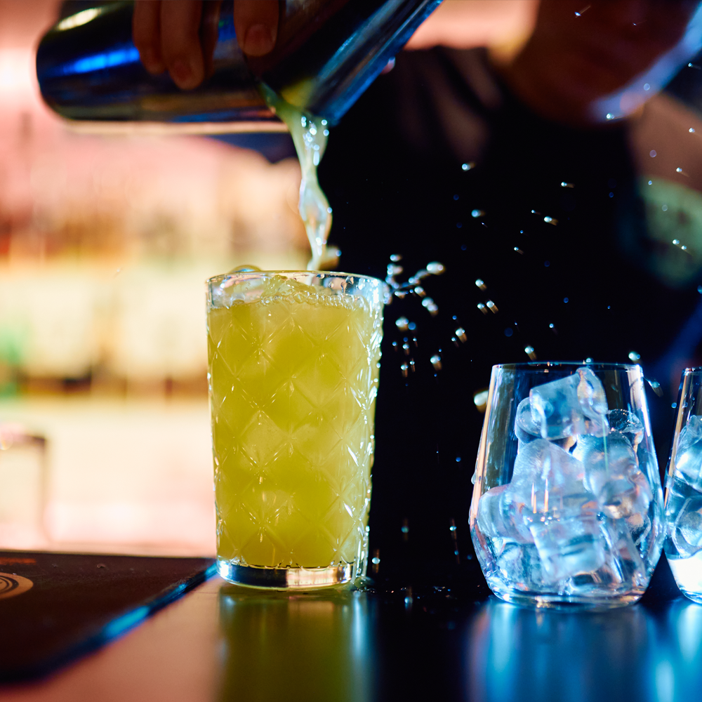 Bartender kaataa keltaisen juoman korkeasta lasista, vieressä on jääpaloja ja muuta juomaa.