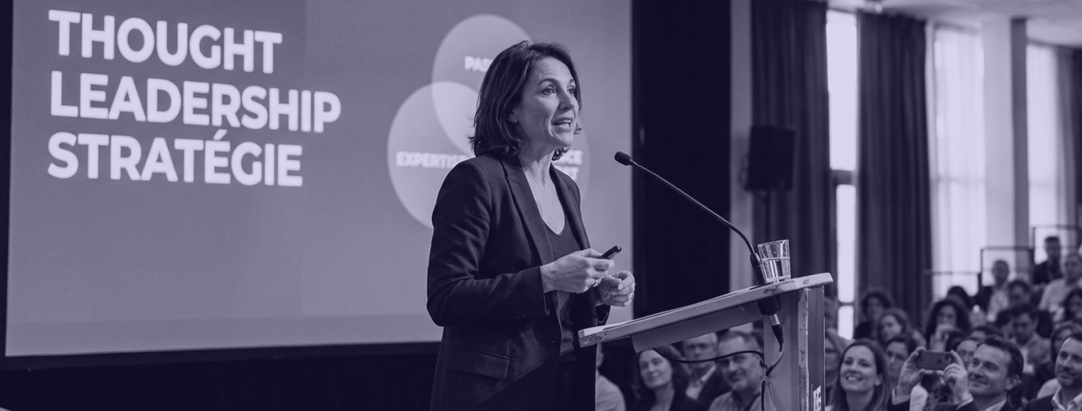Conférencière présentant une intervention sur le leadership et la stratégie derrière un pupitre, face à un public attentif lors d’un événement professionnel.