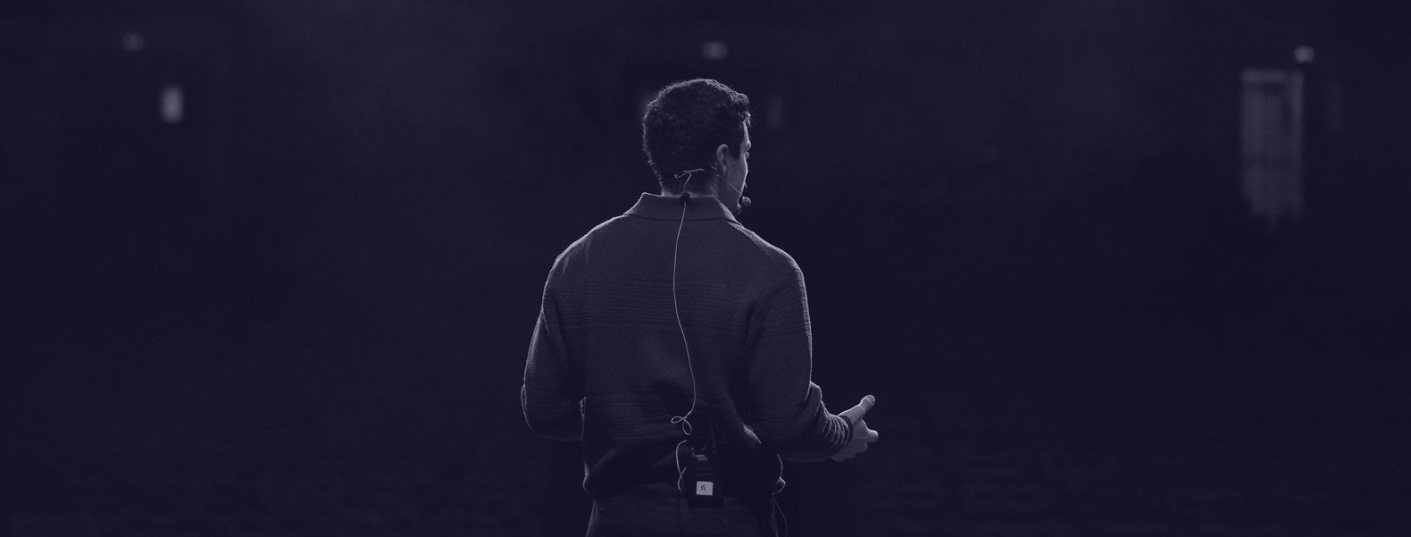 Personne vue de dos sur scène, portant un micro casque, s’adressant à un public dans une salle sombre éclairée par des lumières tamisées.