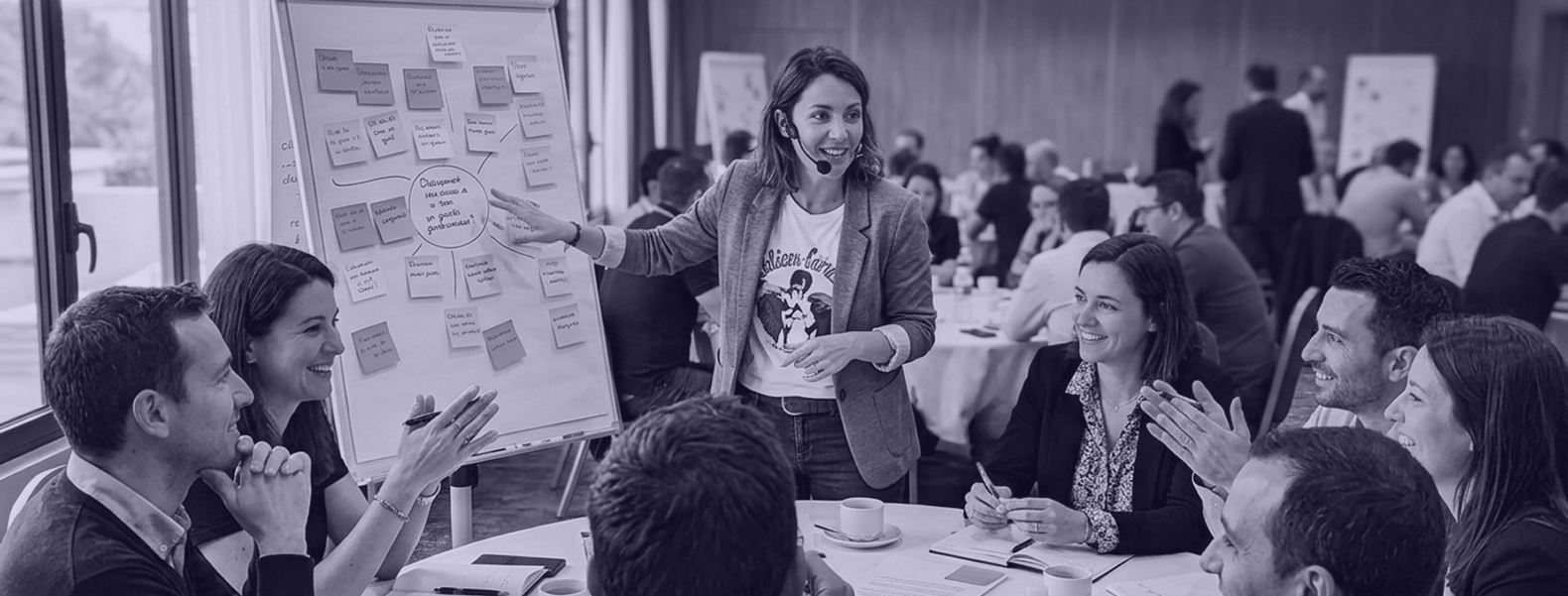 Animatrice portant un micro casque présentant un tableau de post-it lors d’un atelier collaboratif, entourée de participants assis qui échangent et applaudissent.