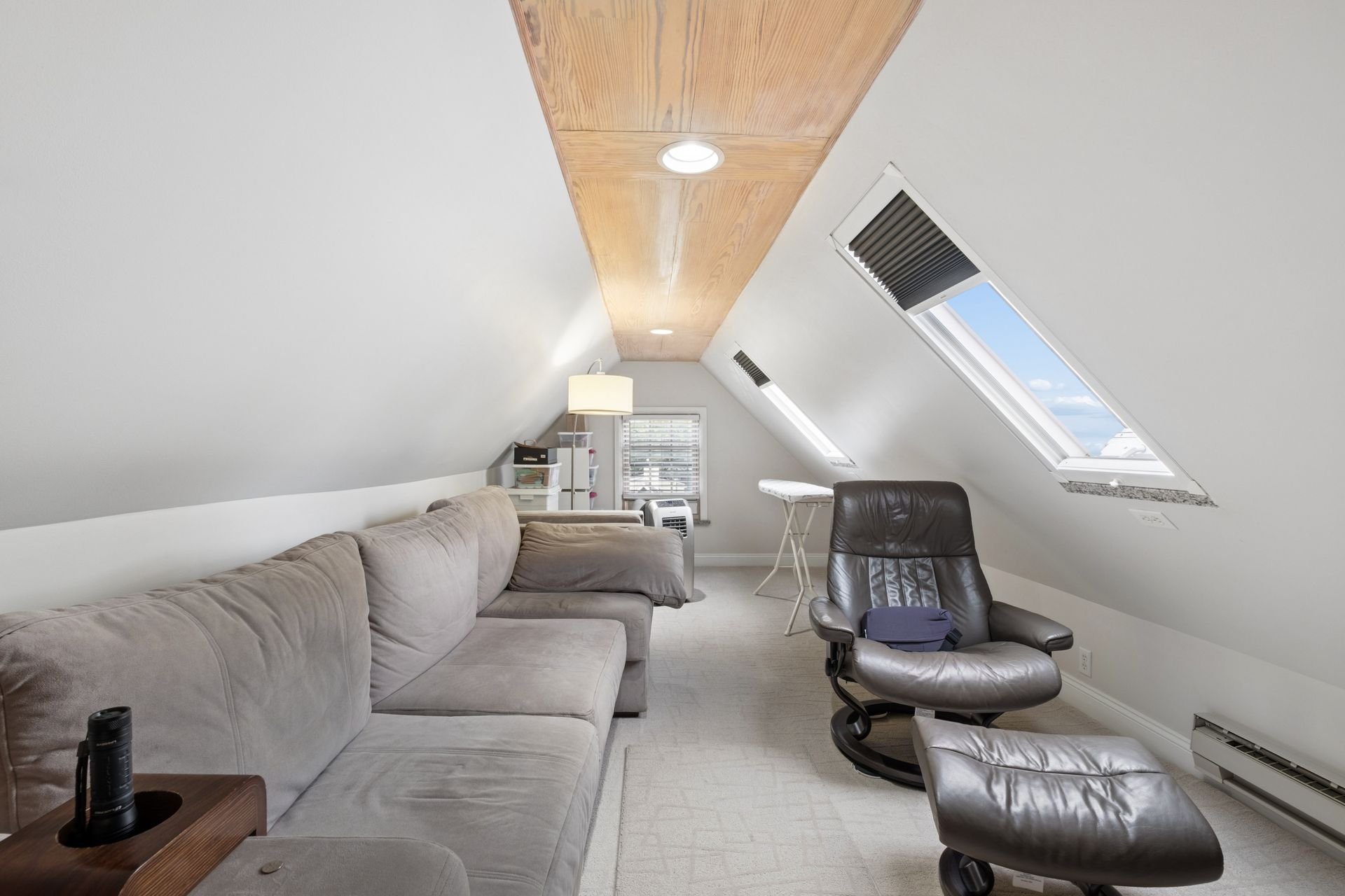 Living room with beige sofa, black leather chair, white window blinds, skylight windows, ceiling wood panel with recessed lights, small side table with binoculars, white lamp, indoor plants, and window with view of blue sky and clouds.