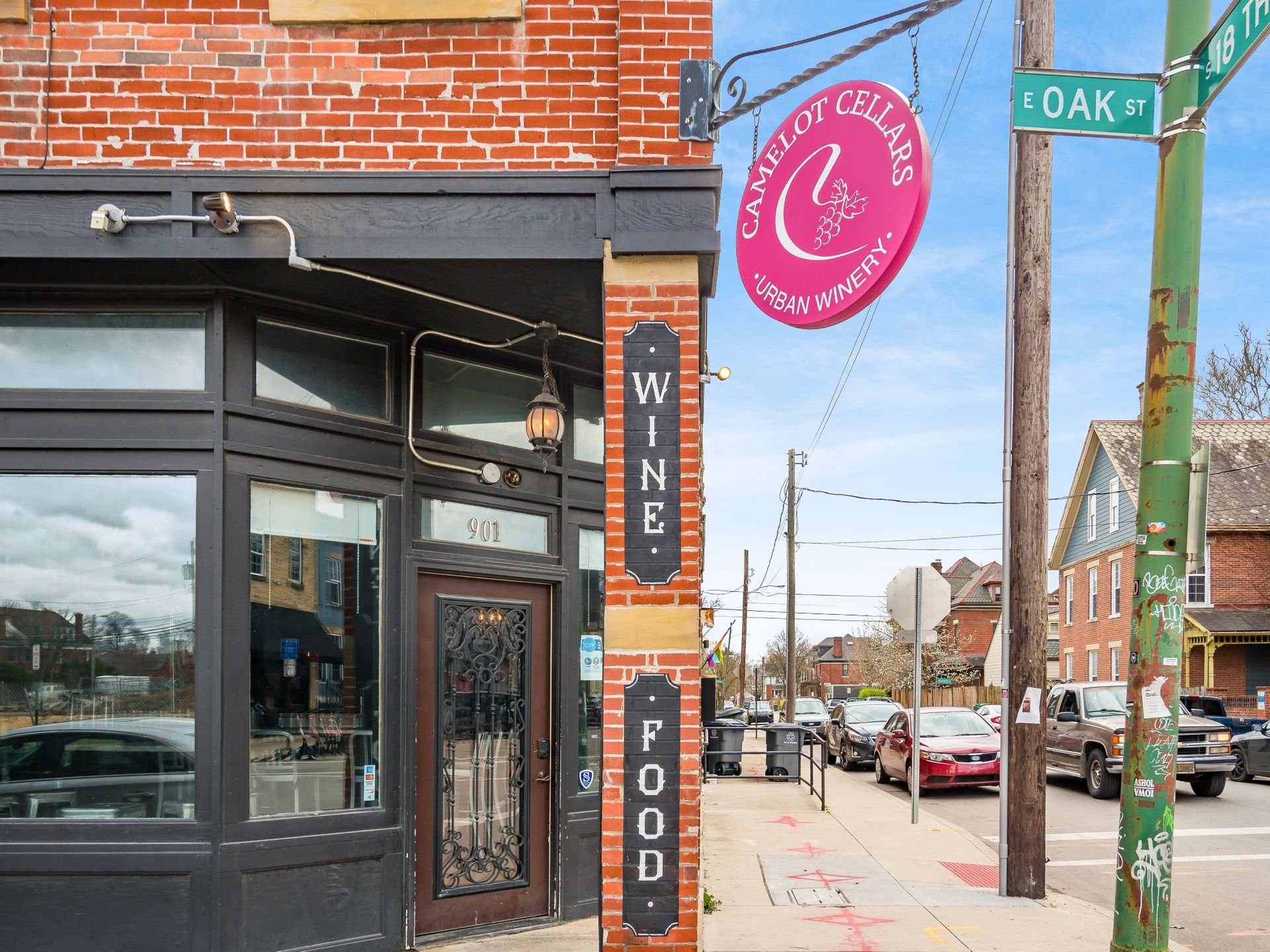 Street view of an urban winery named Camelot Cellars, with a red sign, brick exterior, and "WINE FOOD" displayed vertically on the wall. Located on the corner of E Oak St and another street, with cars and residential buildings in the background.