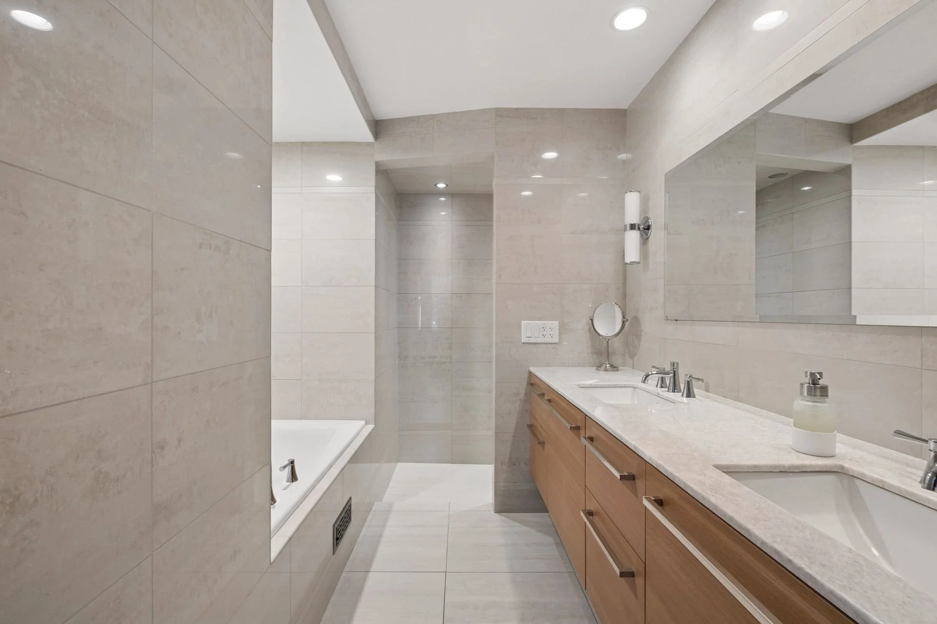 Modern bathroom with beige tiled walls and ceiling, wooden vanity with marble countertop, mirror, wall-mounted light fixture, and bathroom accessories.